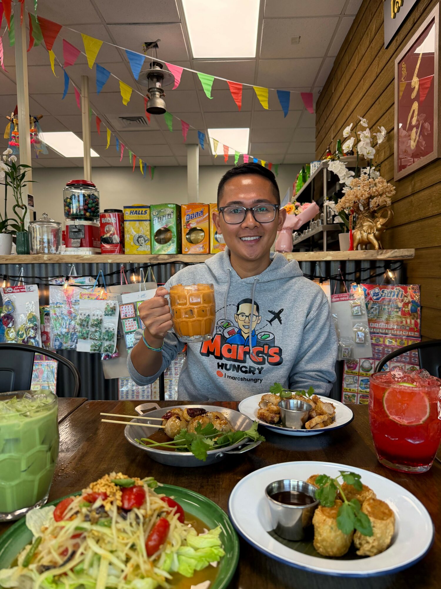 Smiling person wearing glasses and a gray hoodie, holding a mug, seated at a table with food and drinks, decorated room.