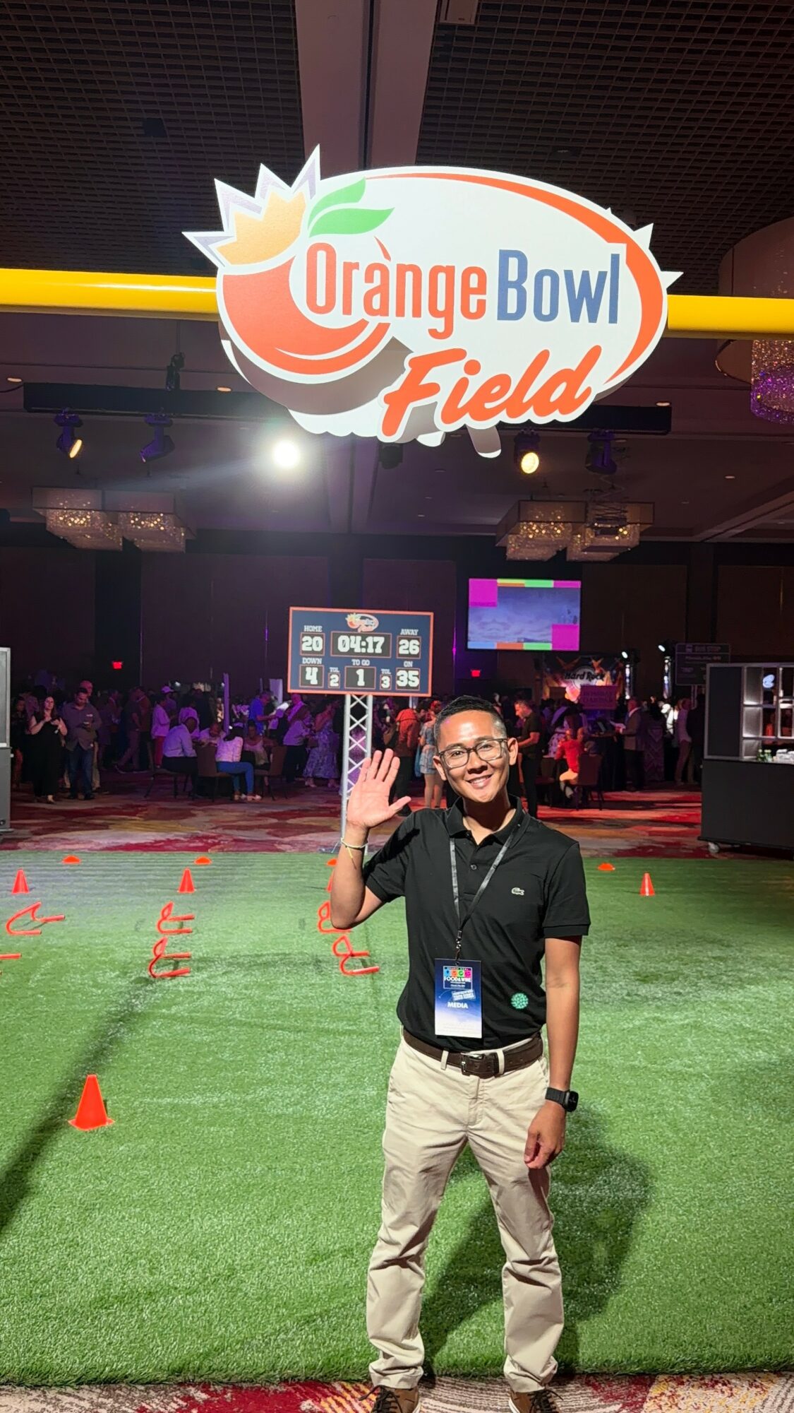 Person standing on green field with orange cones, waving, in front of Orange Bowl Field sign, indoor event space.