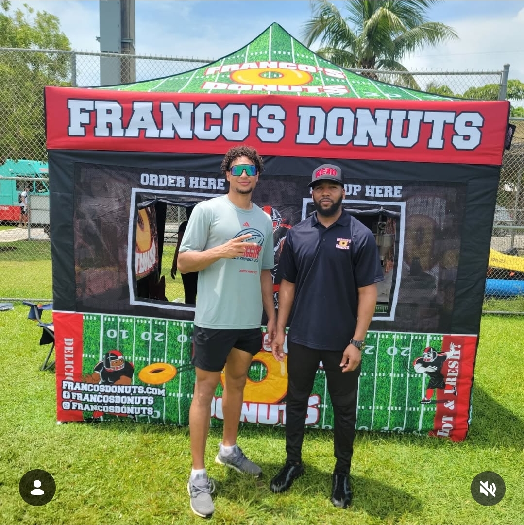 Two men standing in front of a red and green tent with 'FRANCO'S DONUTS' sign, holding drinks, outdoors on grass.