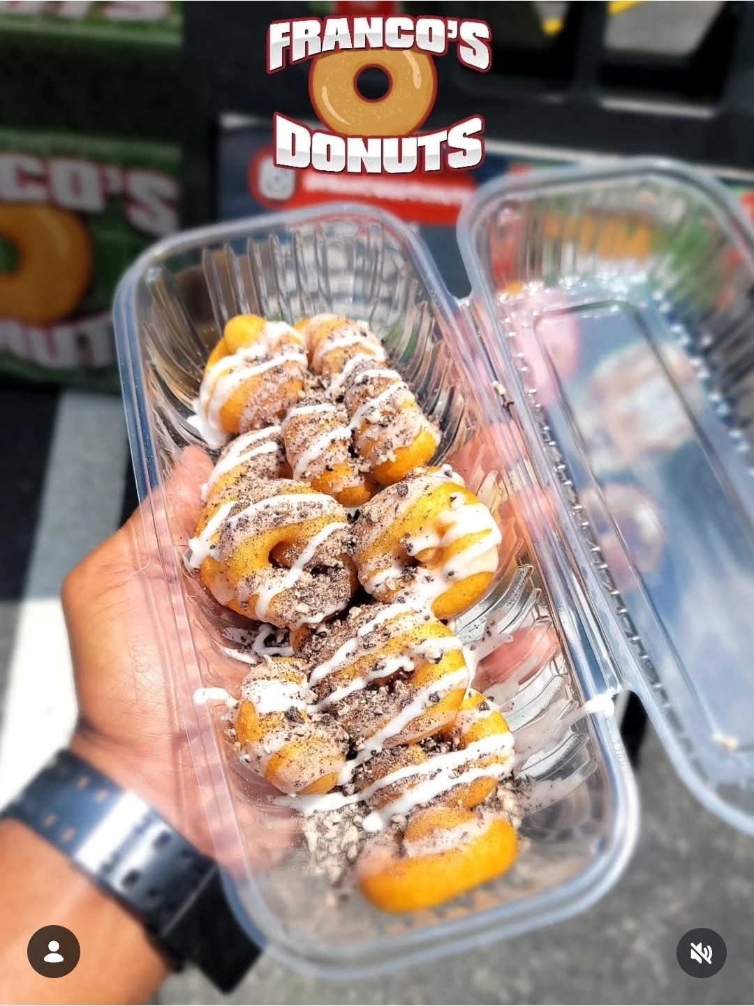 Hand holding a clear plastic container with donuts topped with icing and powdered sugar, in front of a donut box.