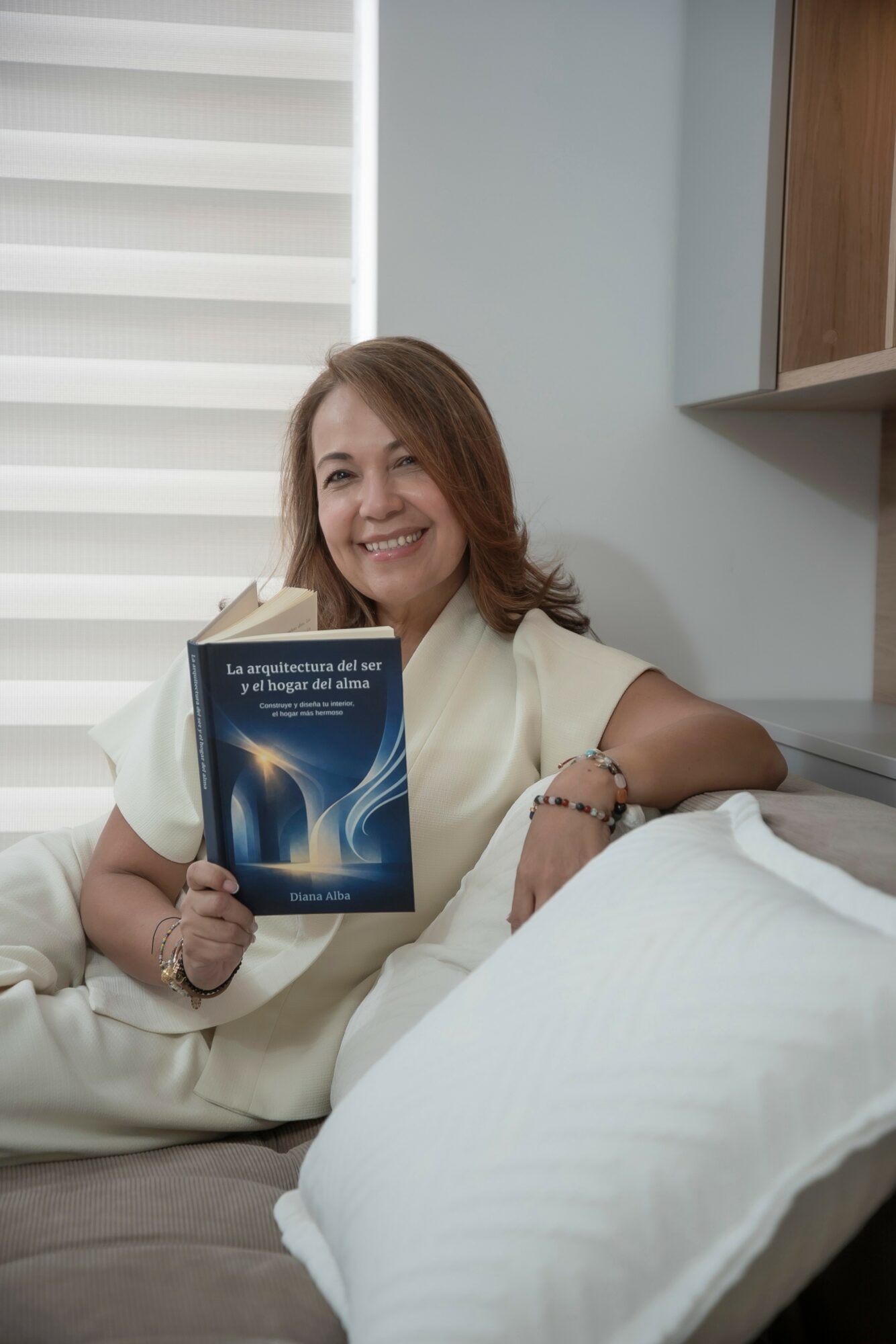 Woman smiling and holding a book while sitting on a bed in a room with white walls and wooden shelves.