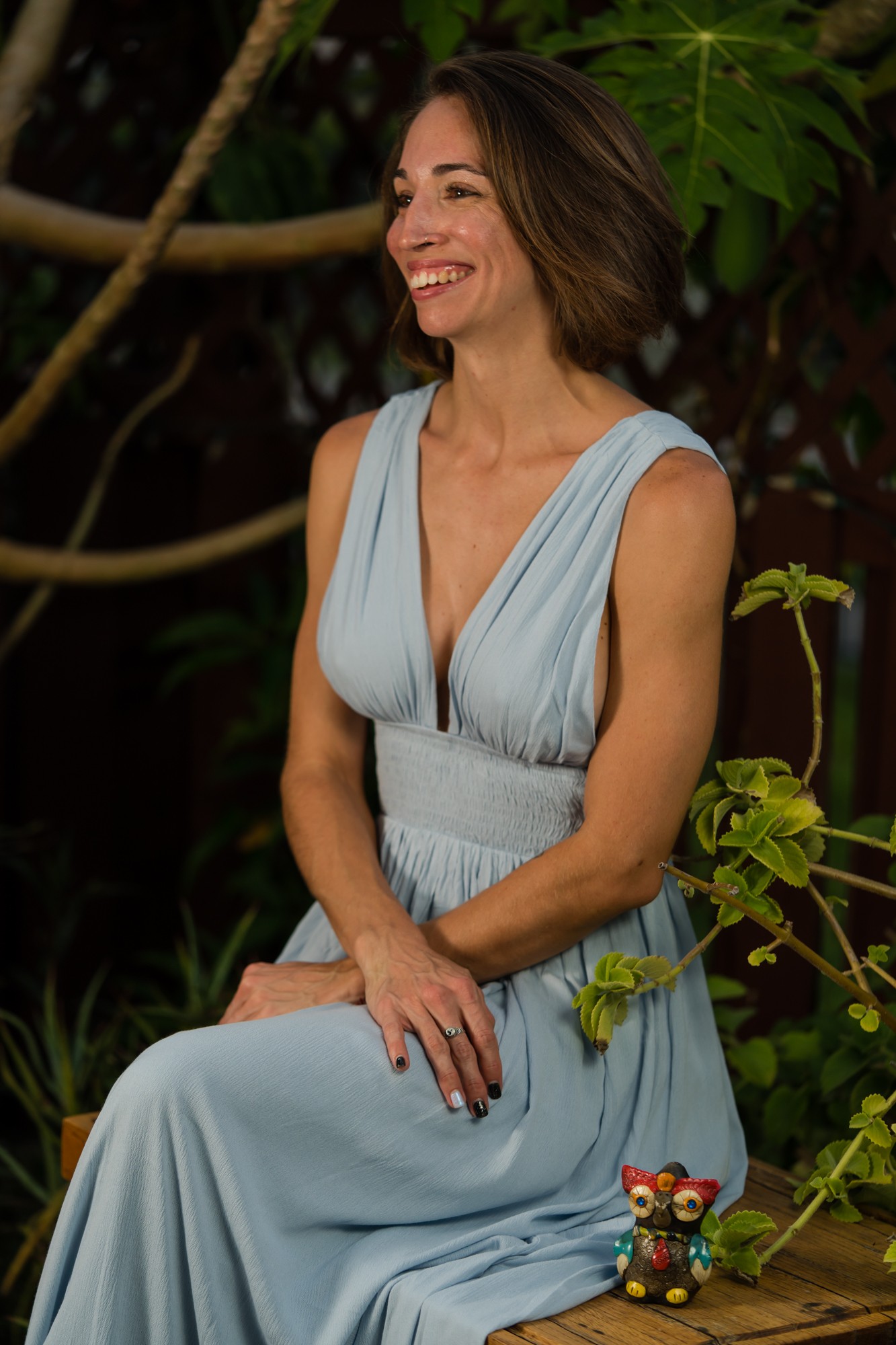 Woman in light blue dress sitting outdoors, smiling, surrounded by green plants and foliage.