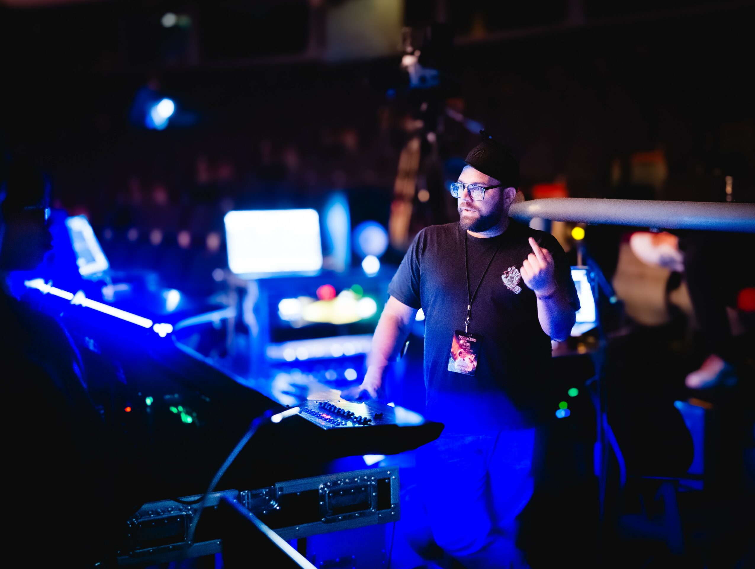 DJ wearing glasses and a beanie, operating equipment in a dark, colorful club setting with screens and lights.