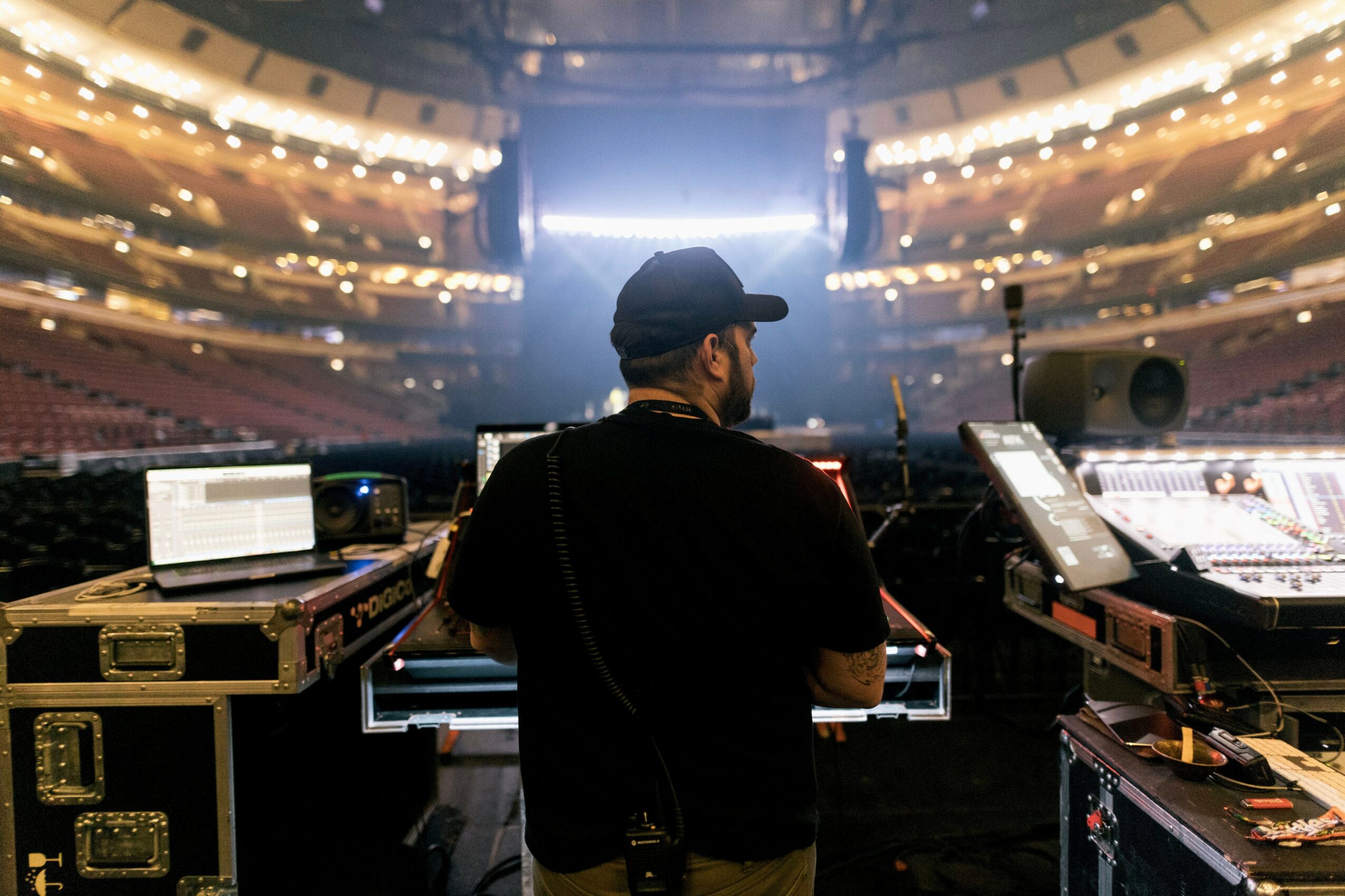 Person wearing a cap working at a DJ booth in a large, empty auditorium with bright lights and multiple screens.
