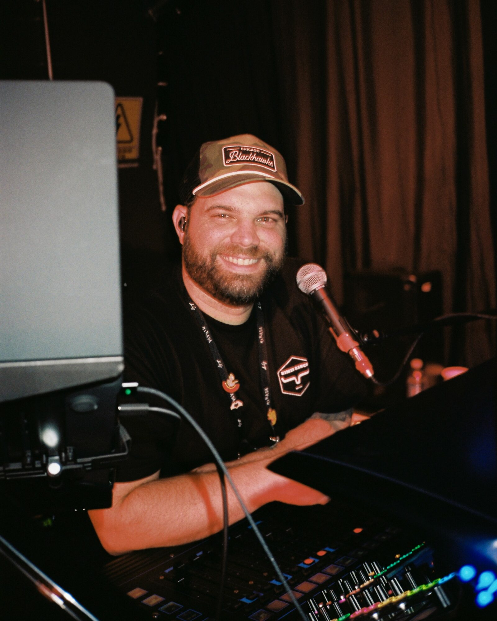 Smiling man with a beard wearing a cap, sitting at a DJ setup with a microphone in a dimly lit room.