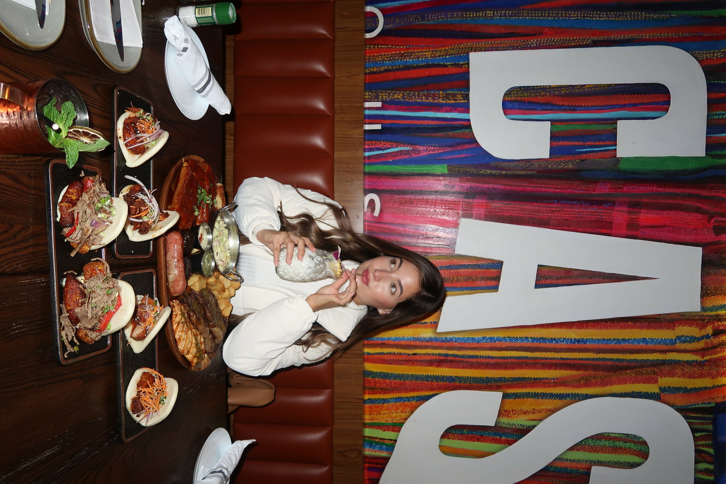 Woman with long hair in white top sitting at table with food and drinks, colorful striped backdrop behind her.