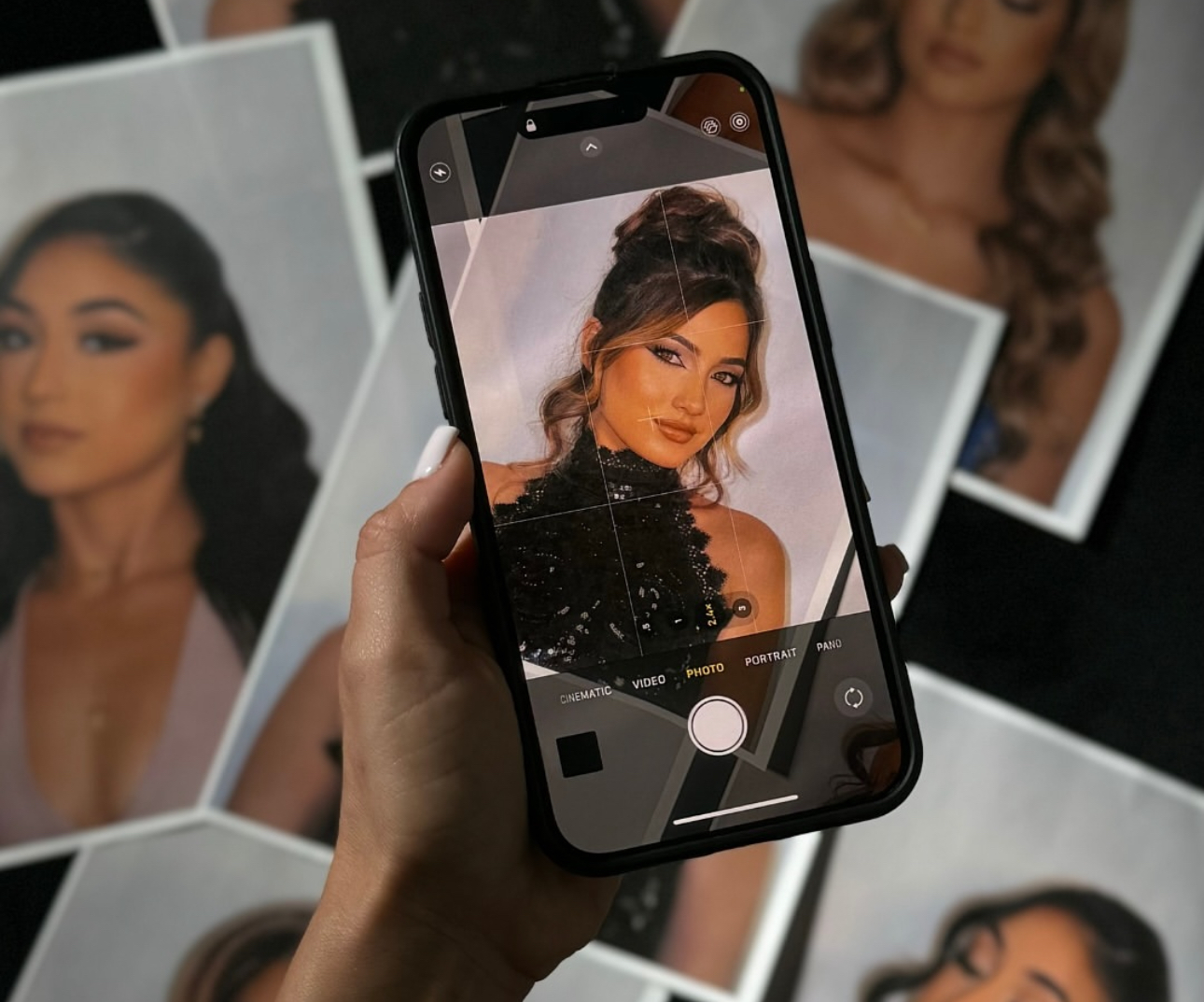 Person holding smartphone taking a photo of a woman with styled hair and makeup, with photos in the background.