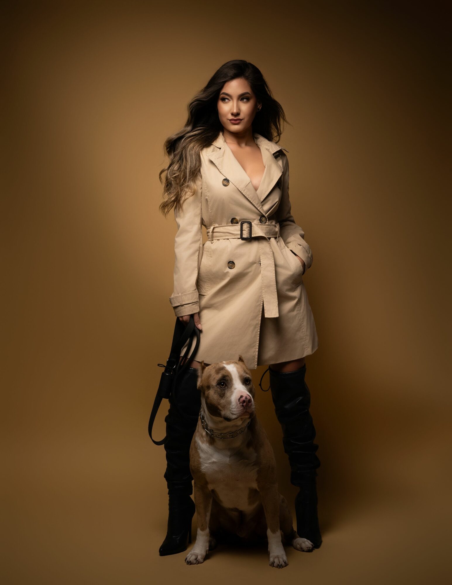 Woman in beige trench coat standing with a dog on a leash against a brown background.