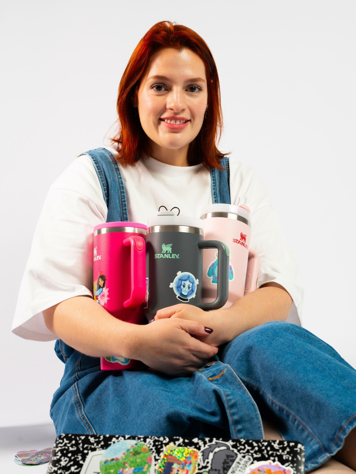 Young woman with red hair holding four colorful mugs, sitting cross-legged, smiling, against a plain white background.