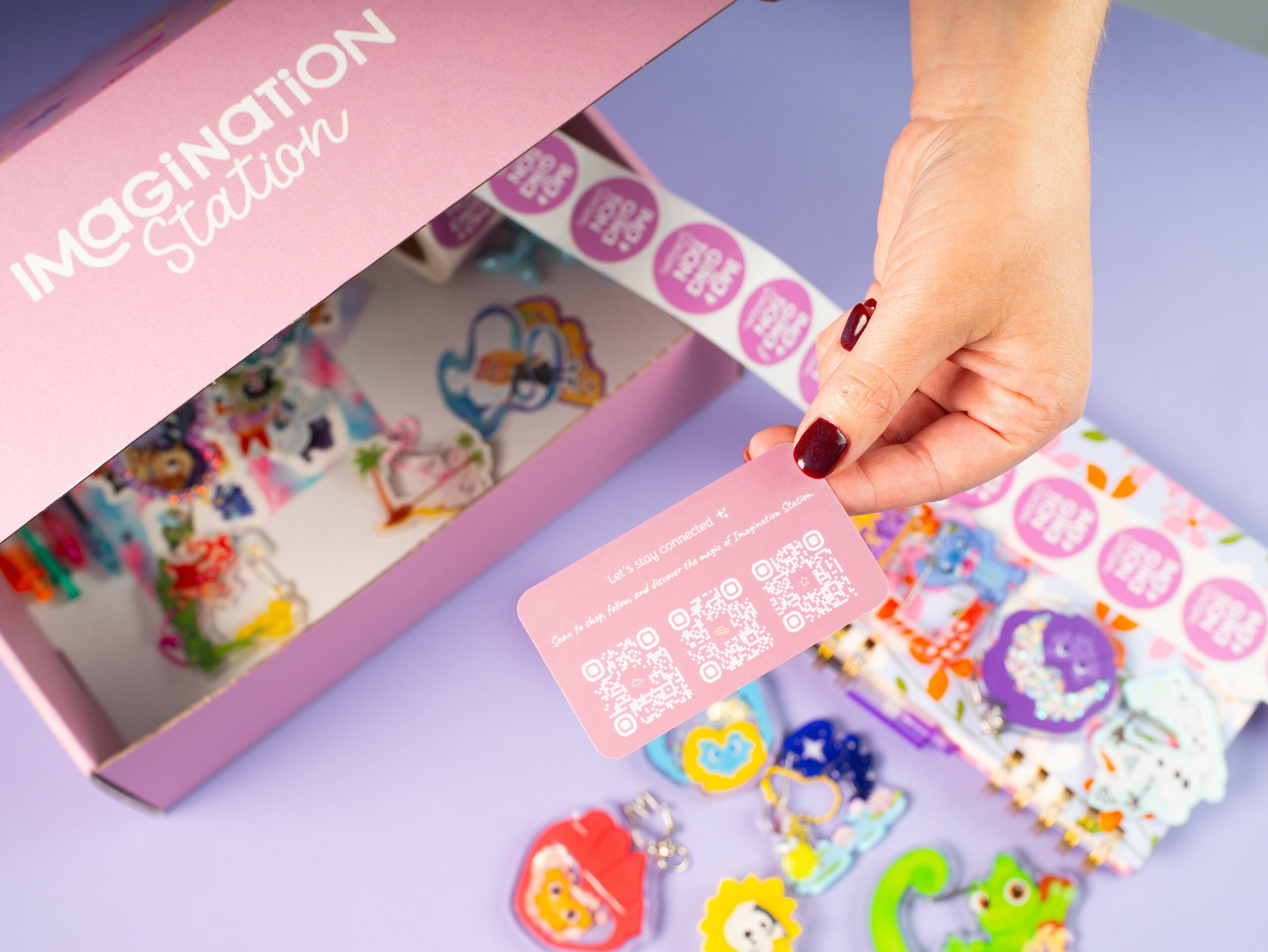 Hand holding a pink card above colorful bracelet pieces on a pink box labeled 'Imagination Station'.