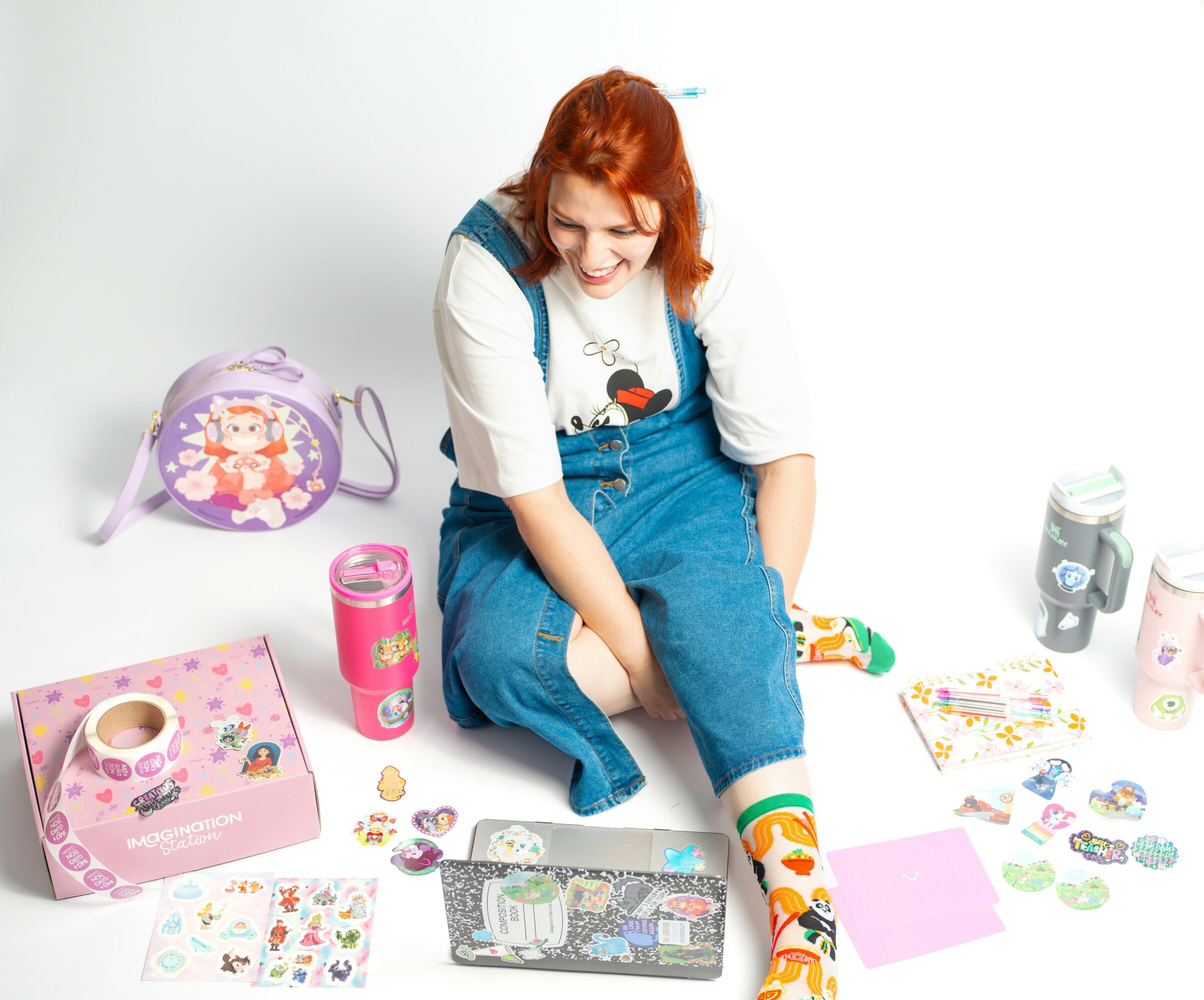 Girl with red hair sitting on floor, surrounded by stickers, toys, and a pink box, smiling and looking down.