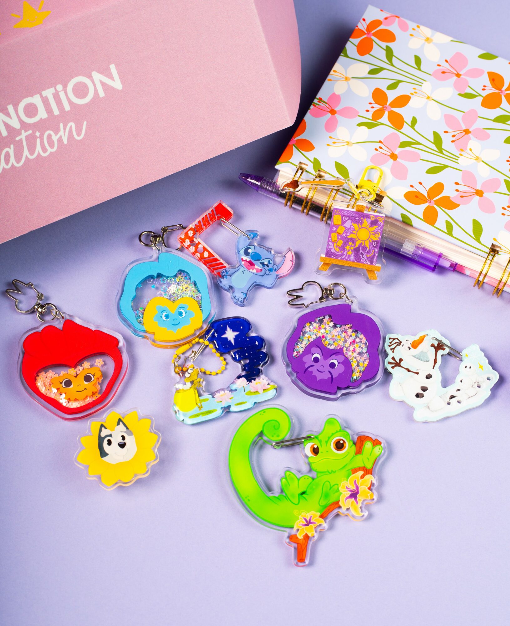 Collection of colorful keychains with animal and character designs on a purple surface, with a pink box and floral notebook nearby.
