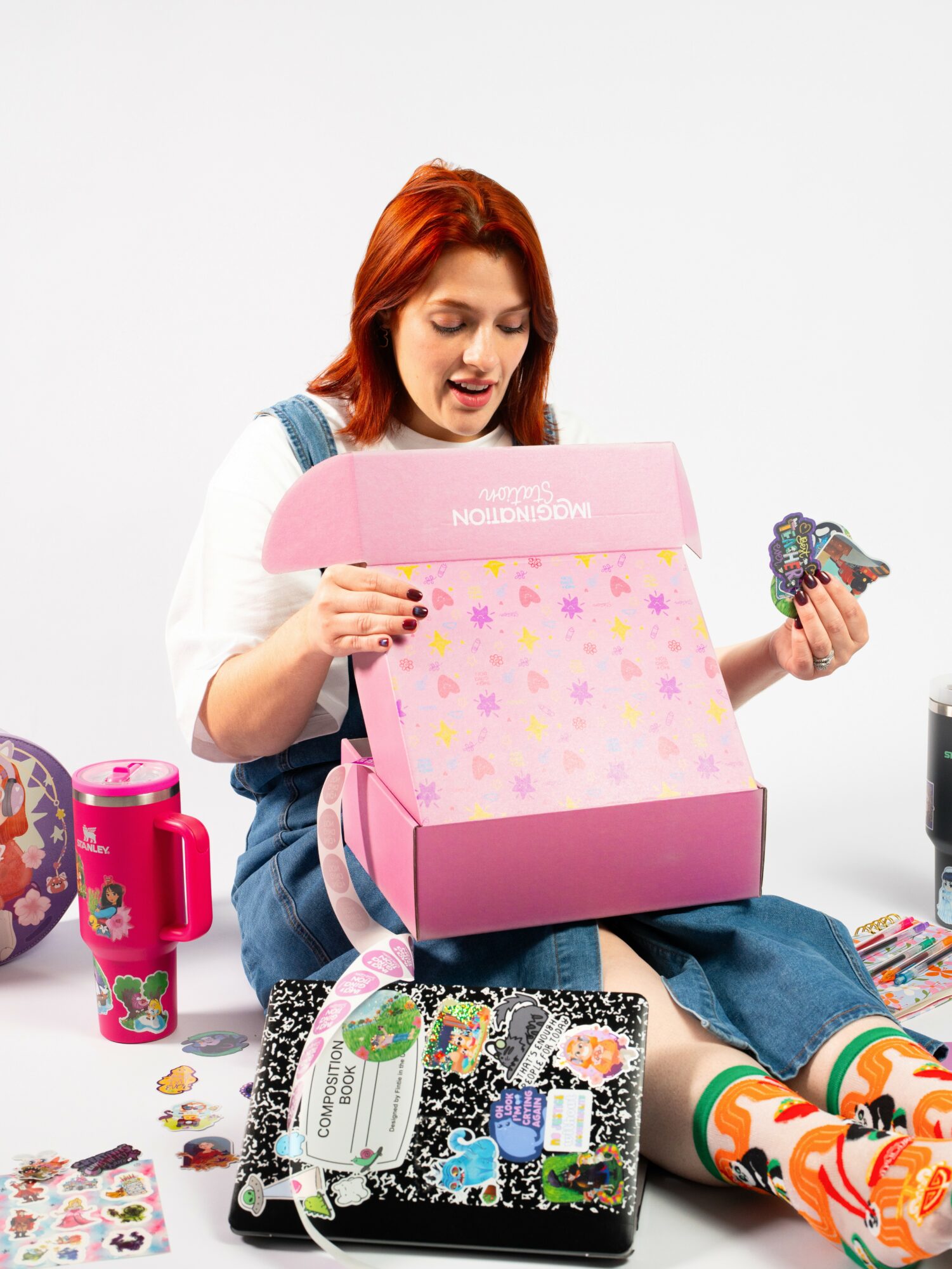 Young woman with red hair opening a pink gift box with colorful stars, surrounded by gifts and wrapping paper.