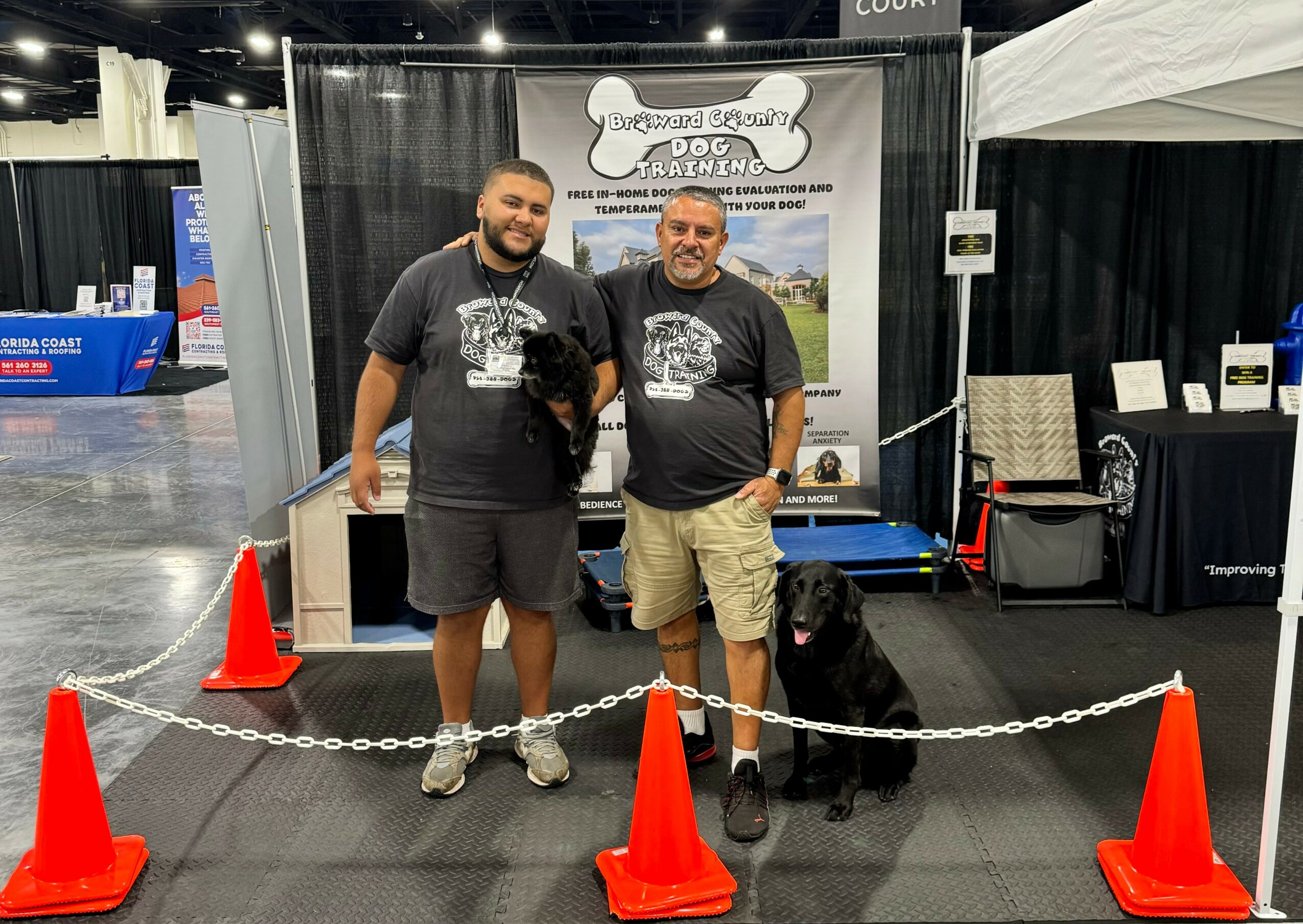 Two men standing behind a chain barrier with a black dog sitting beside them, at an indoor event booth.