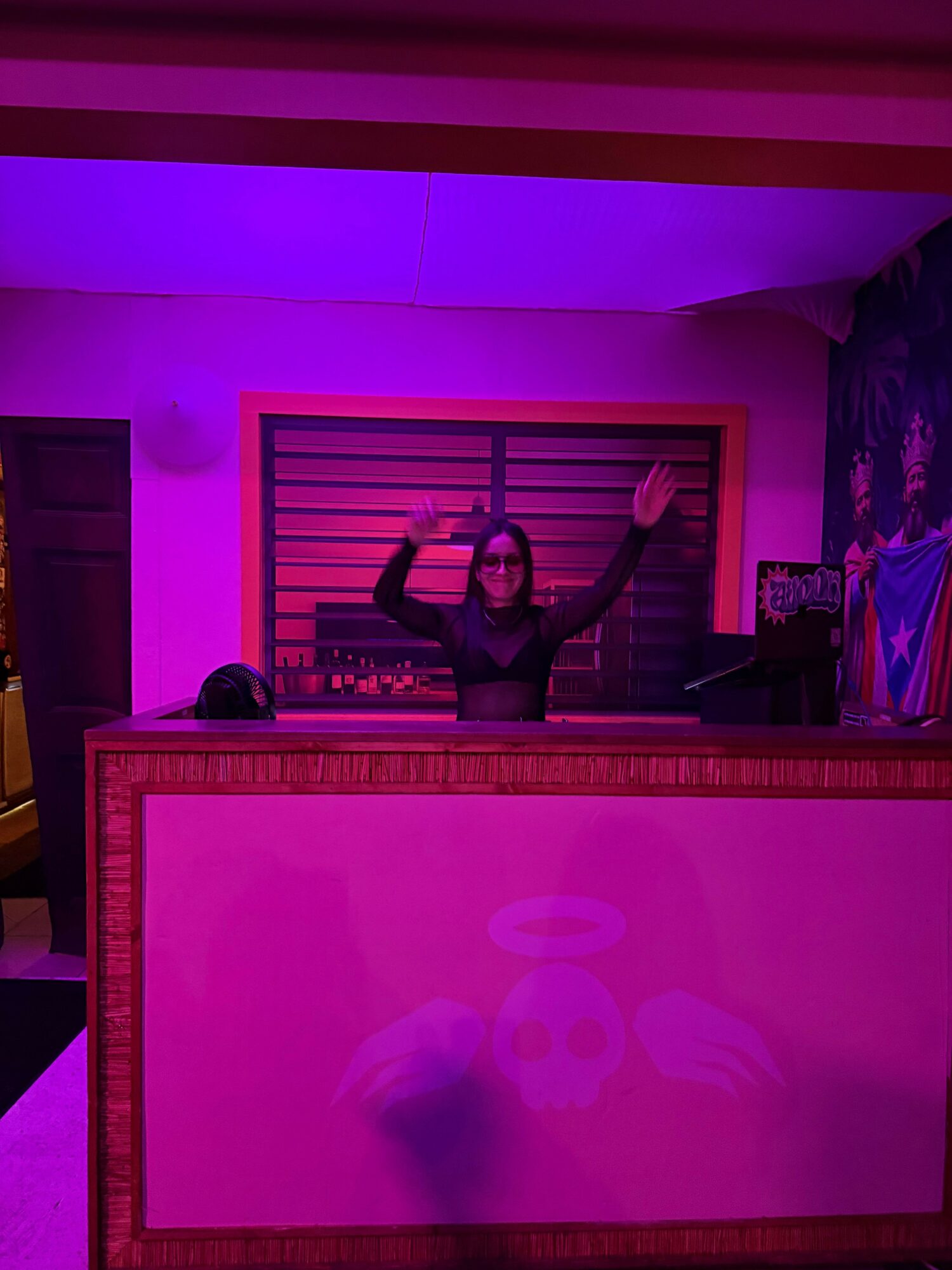Woman with glasses raising arms behind a pink-lit counter in a colorful room.