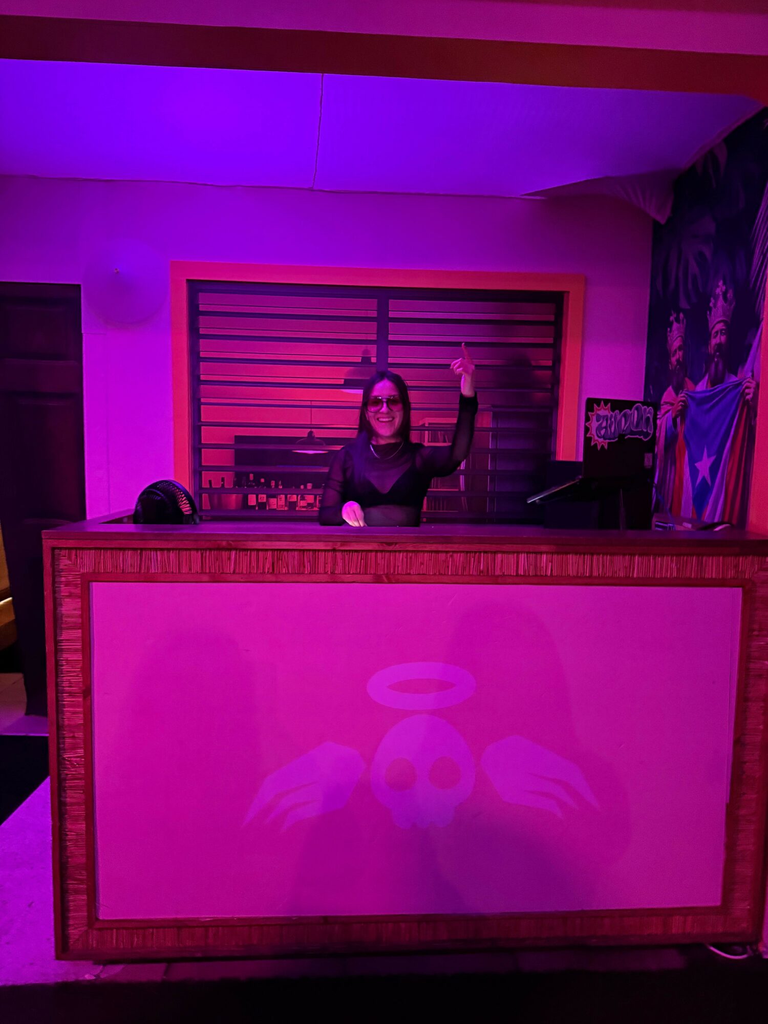 Person standing behind a reception desk, raising one hand, in a room with purple and pink lighting.
