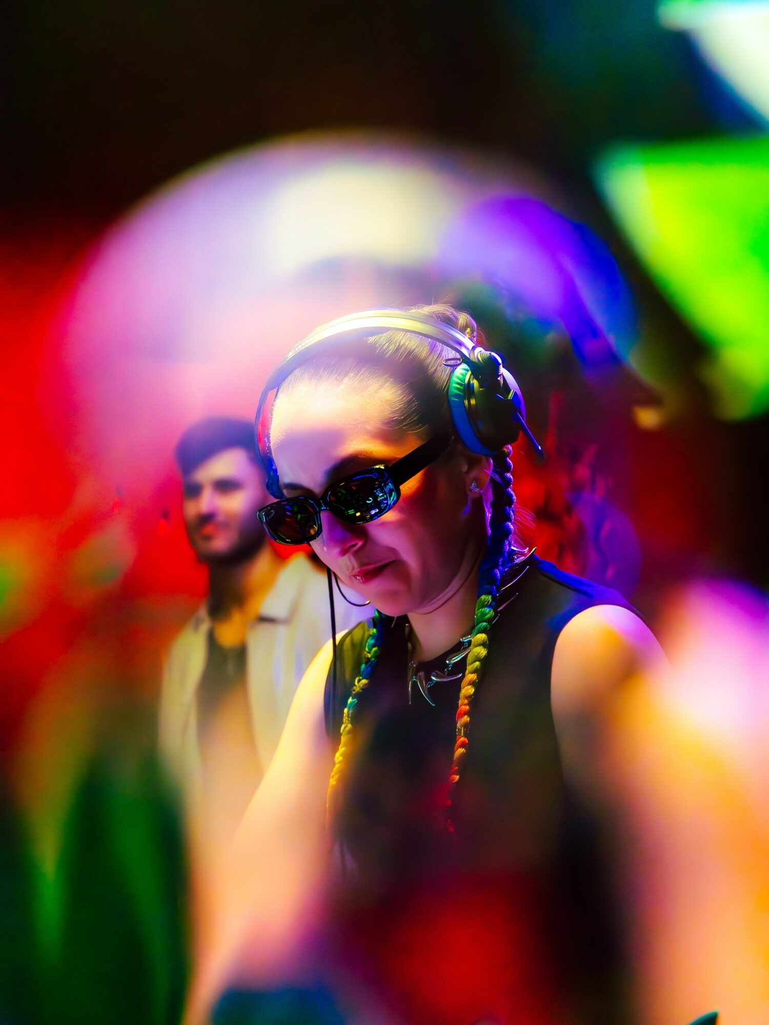 Woman wearing headphones and sunglasses, with colorful blurred background and a man in the background.