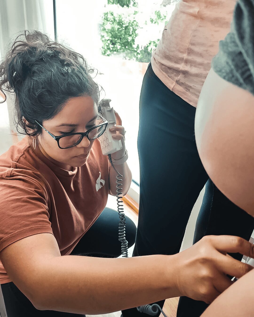 Young woman with glasses and curly hair using a phone, with a pregnant person nearby.