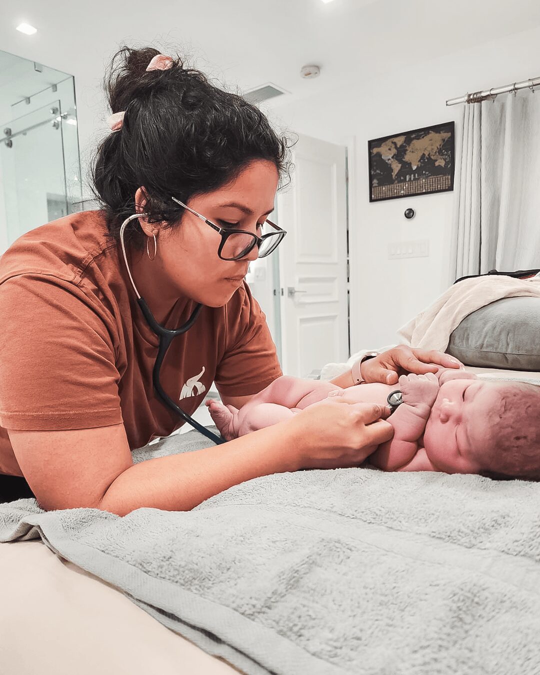 Woman with glasses and stethoscope examining a newborn baby on a bed in a room.