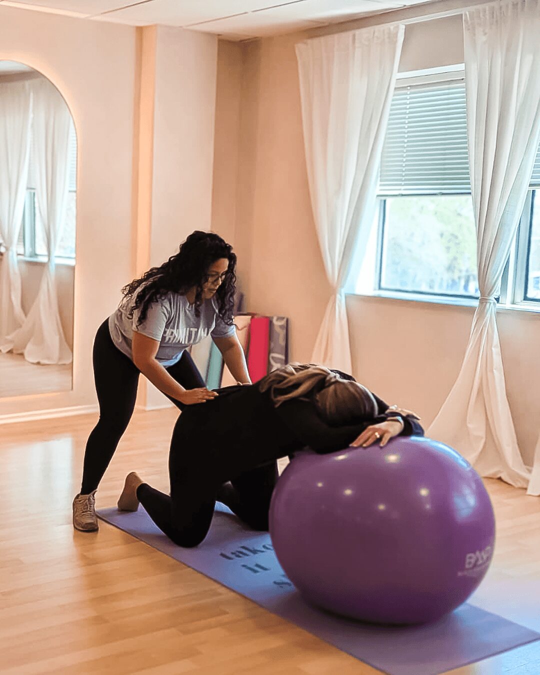 Two women in a room with large windows and white curtains, one kneeling and the other lying on a purple exercise ball.