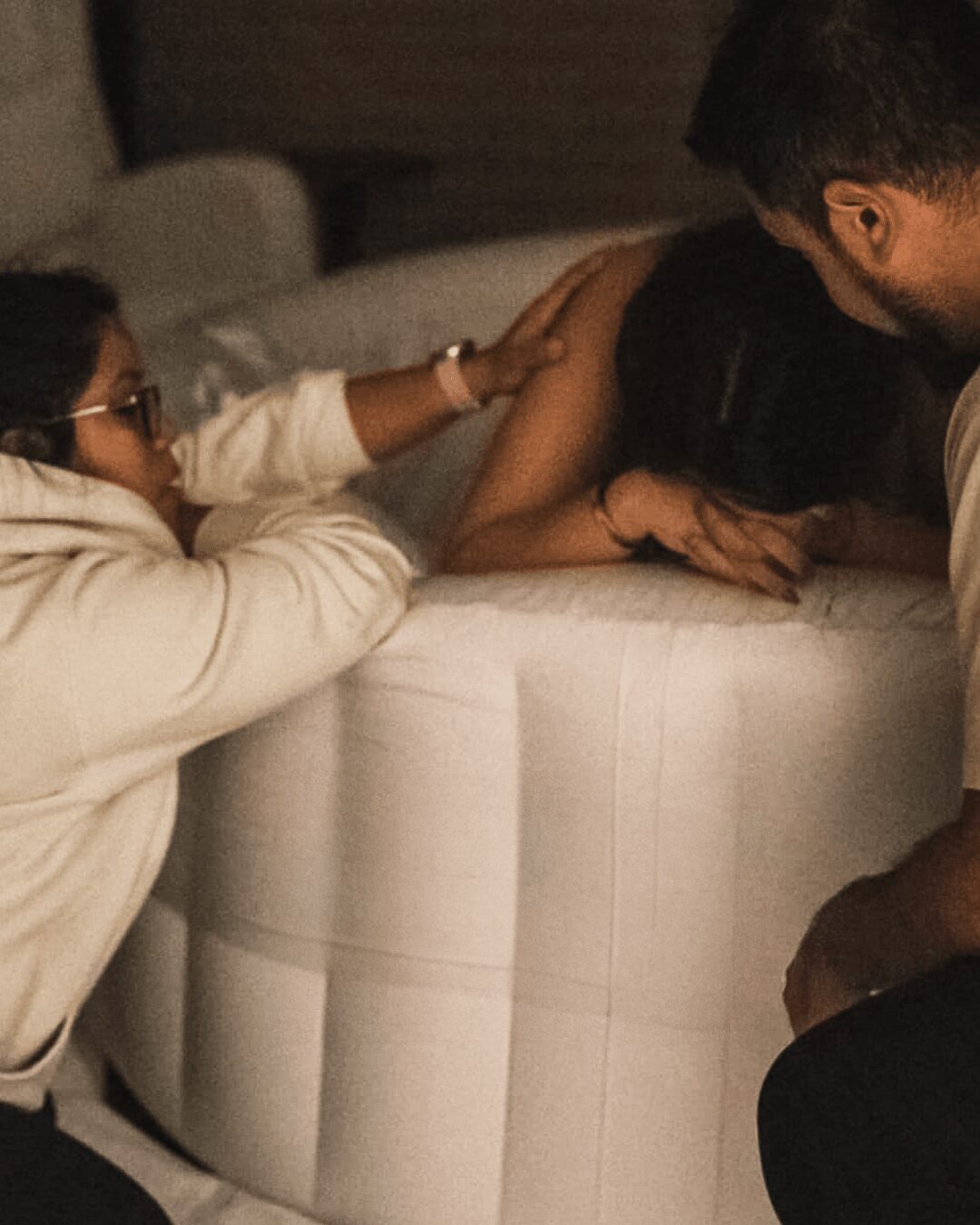 Three people are gathered around a white, rounded piece of furniture, with one person resting their head on it.