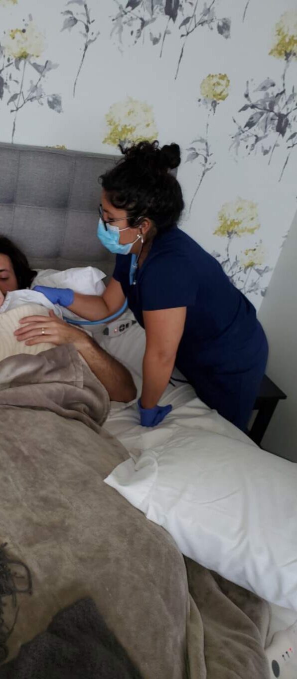 Healthcare worker in blue scrubs and mask tending to a patient in bed.