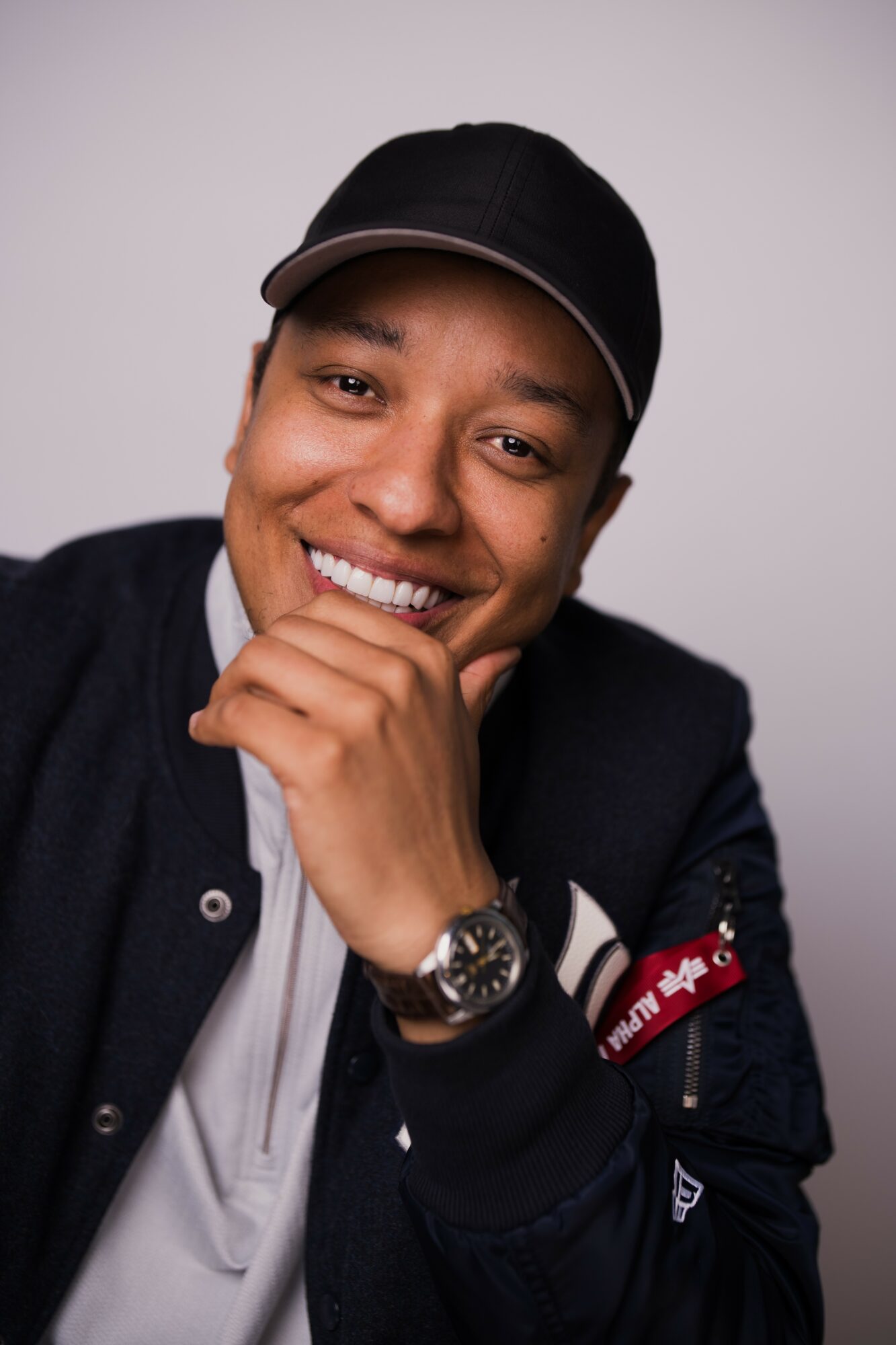 Young man smiling, wearing a black cap and jacket, with hand near chin, against a plain background.