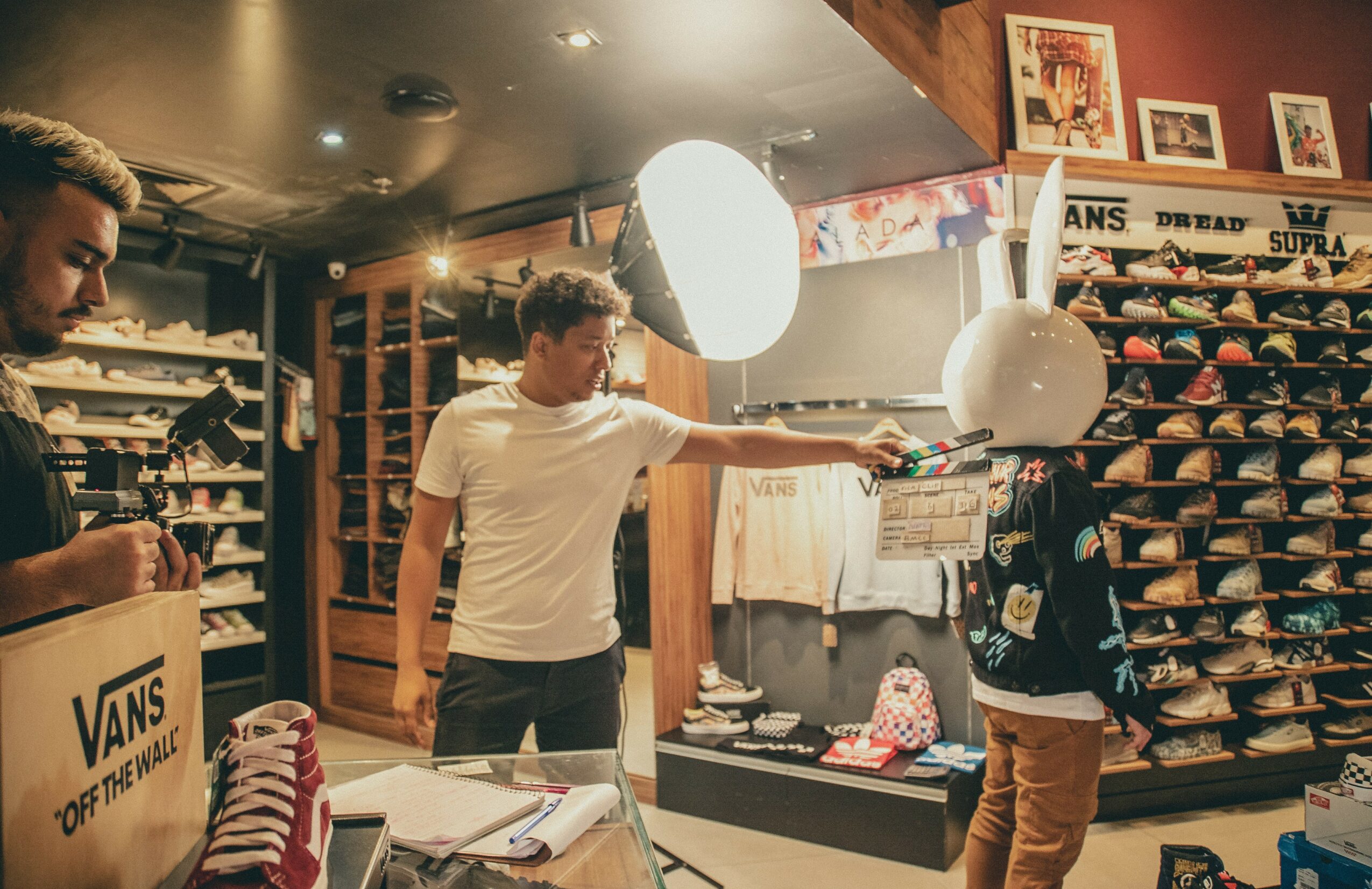 Young man in white shirt reaching for a shoe in a shoe store with shelves of shoes and a large light overhead.