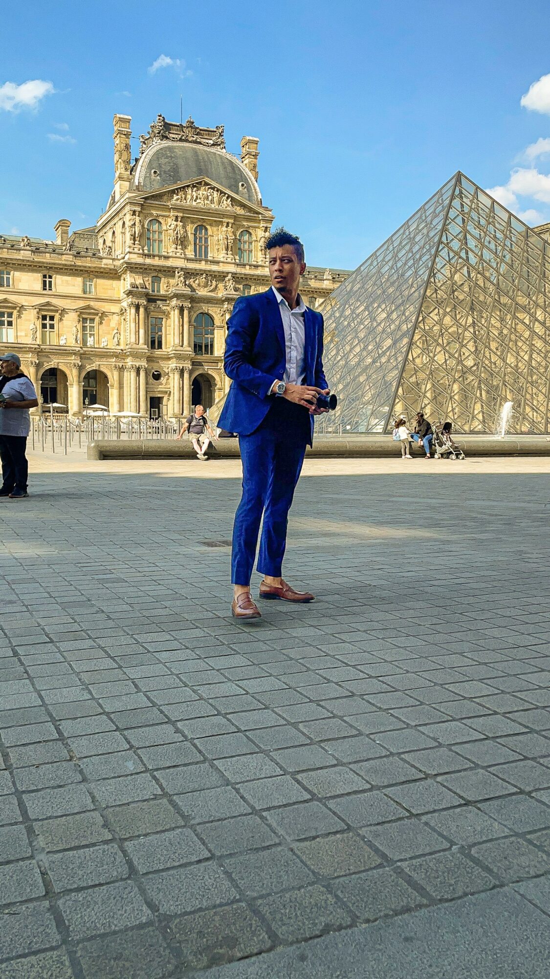 Person in blue suit holding a book standing in front of the Louvre Museum and glass pyramid in Paris.
