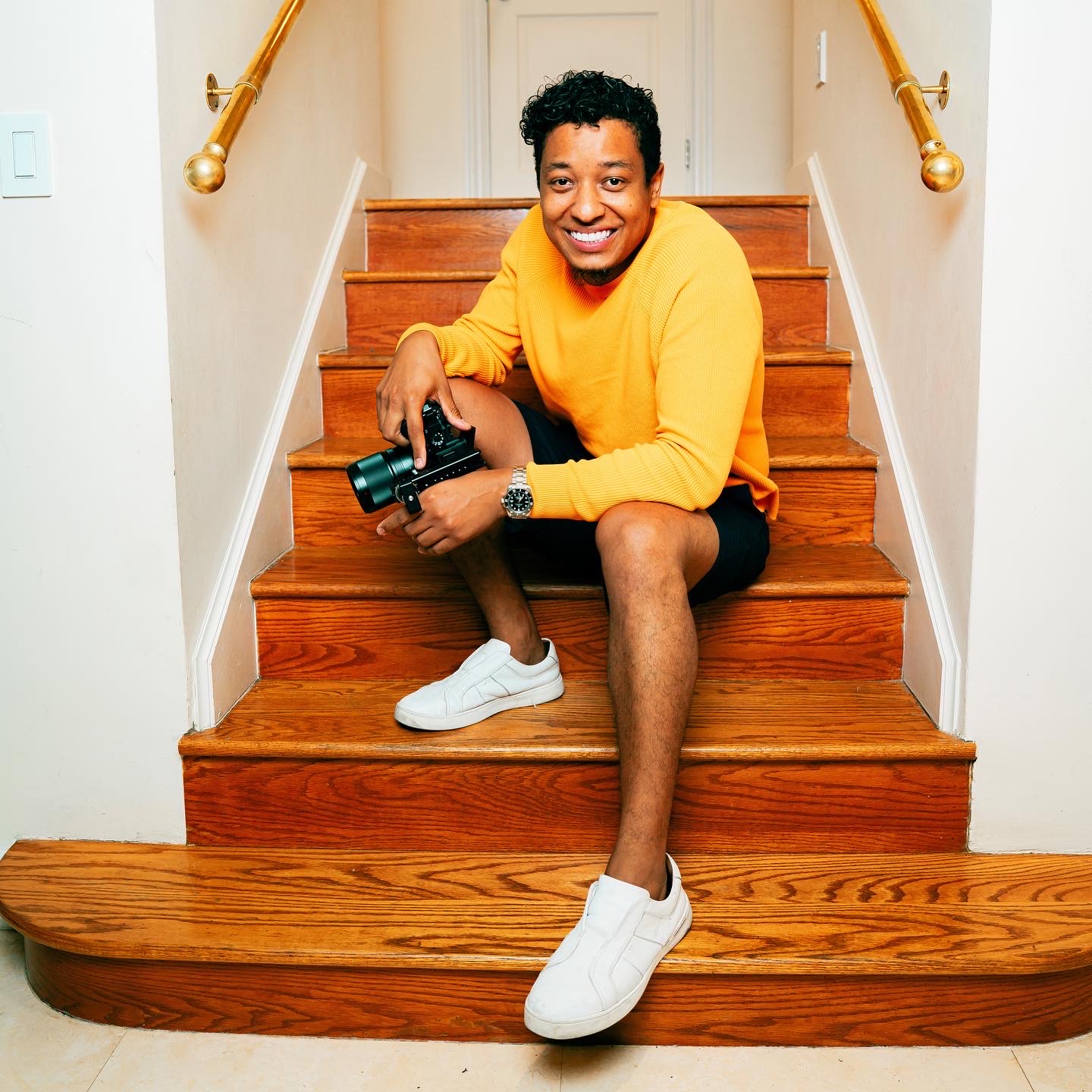 Person sitting on wooden staircase holding a camera, smiling, wearing a yellow shirt, black shorts, and white sneakers.