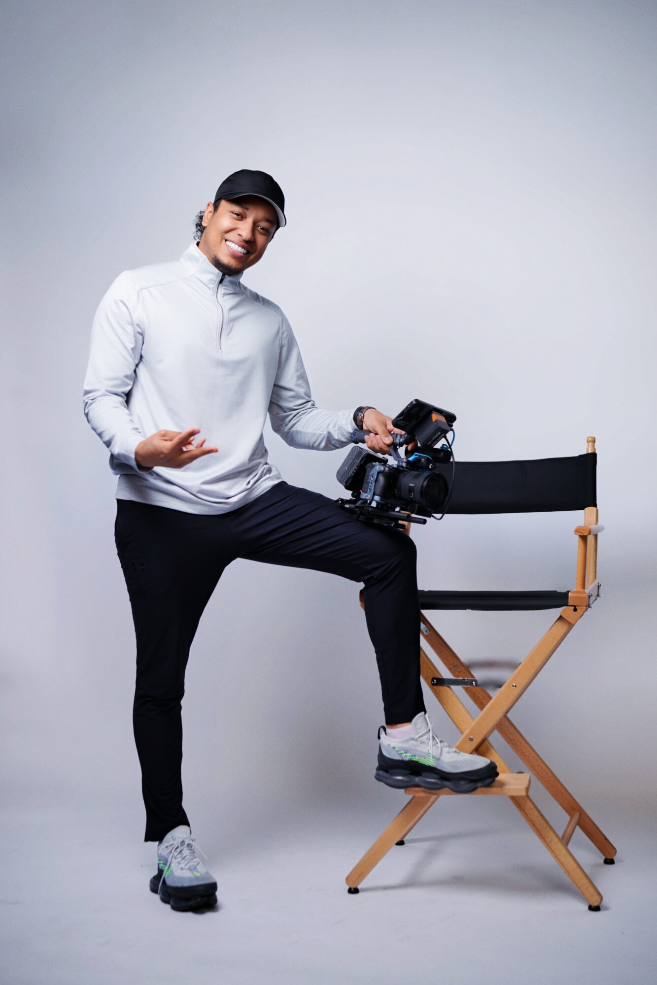 Person standing with one foot on a director's chair, holding a camera, smiling, wearing a cap and casual clothing.