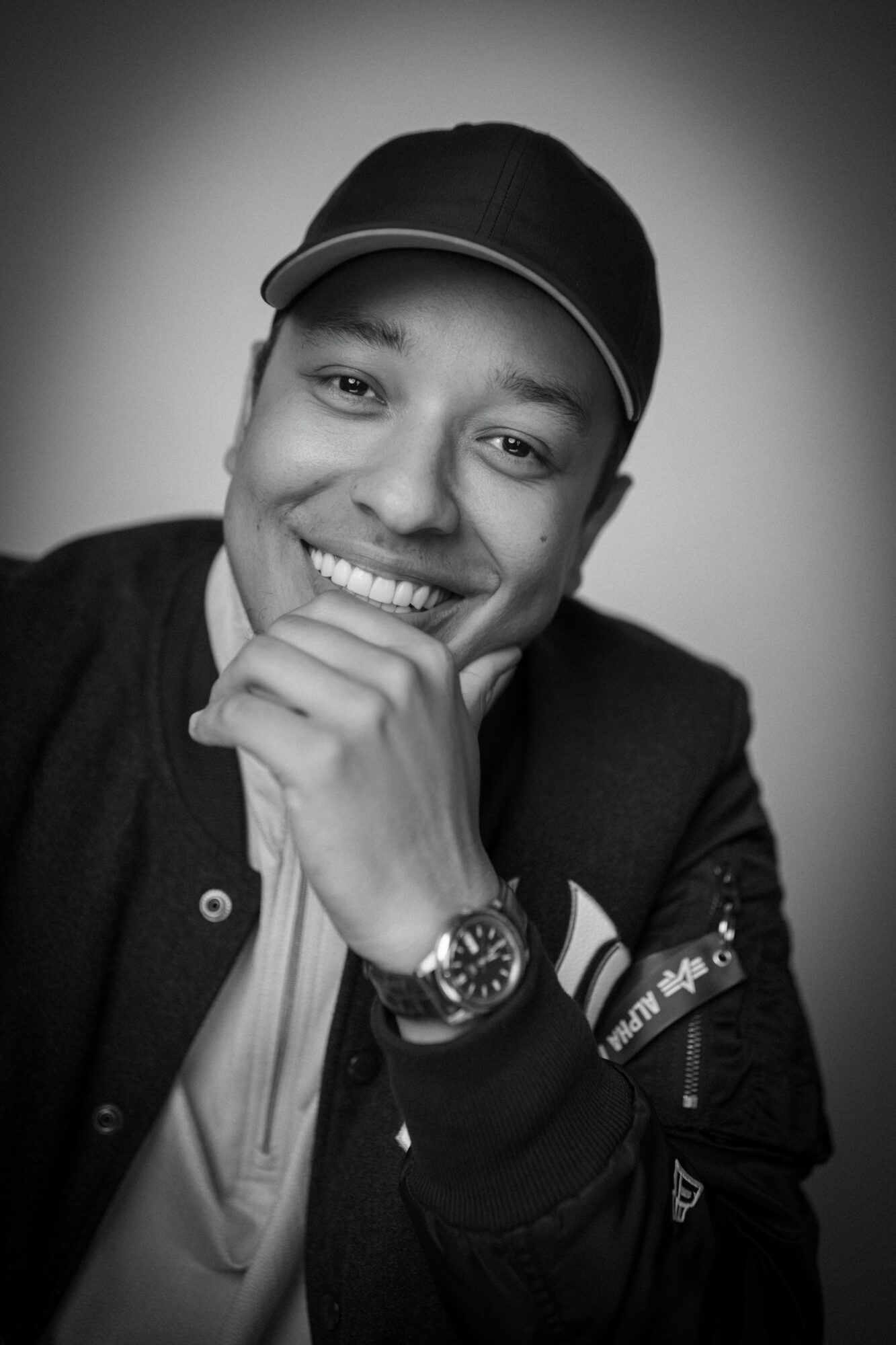 Young man smiling, wearing a cap and jacket, resting his chin on his hand, in black and white.
