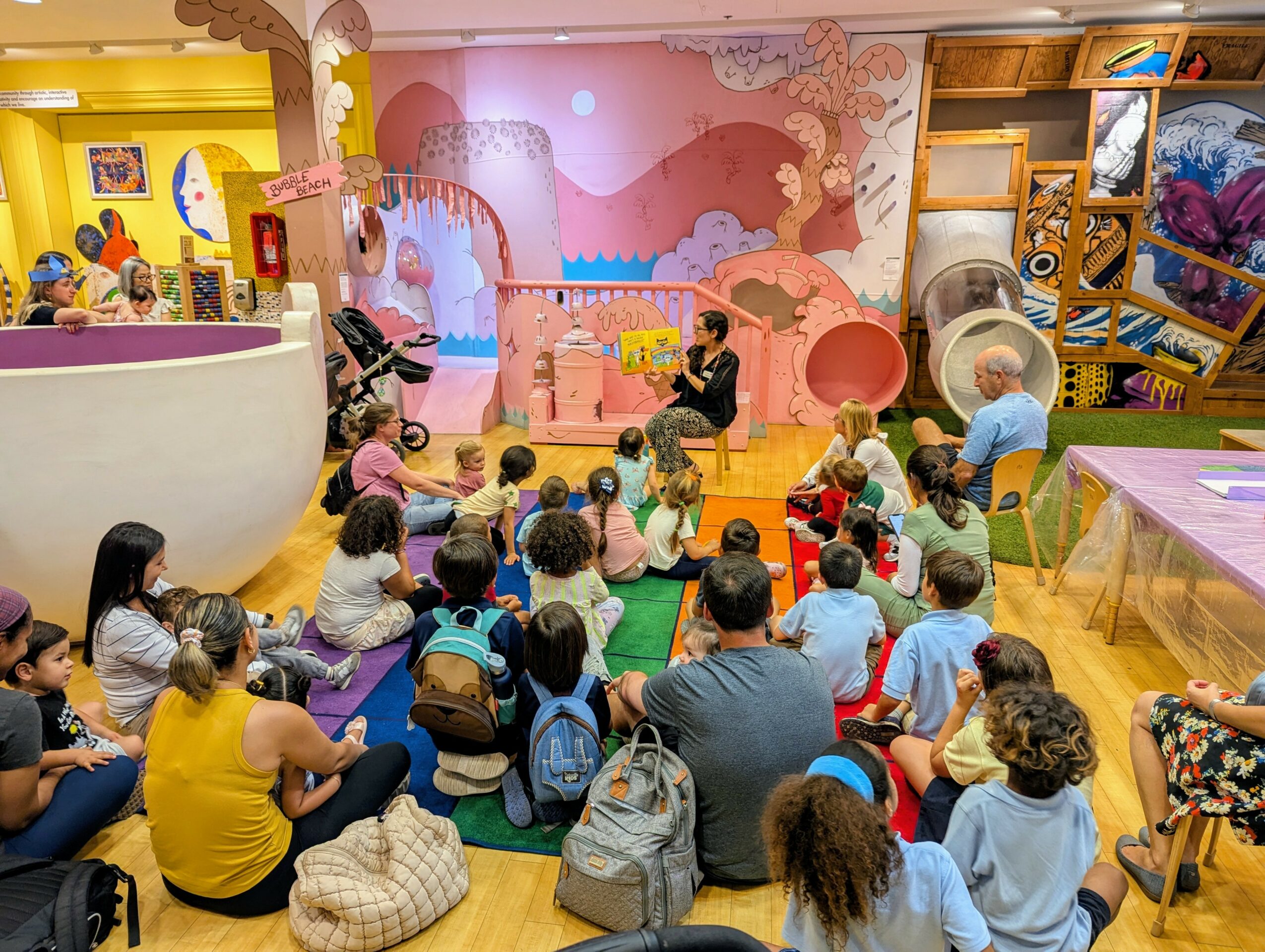Group of children and adults seated on colorful mats in a vibrant indoor play area with pink and yellow walls and playful decorations.