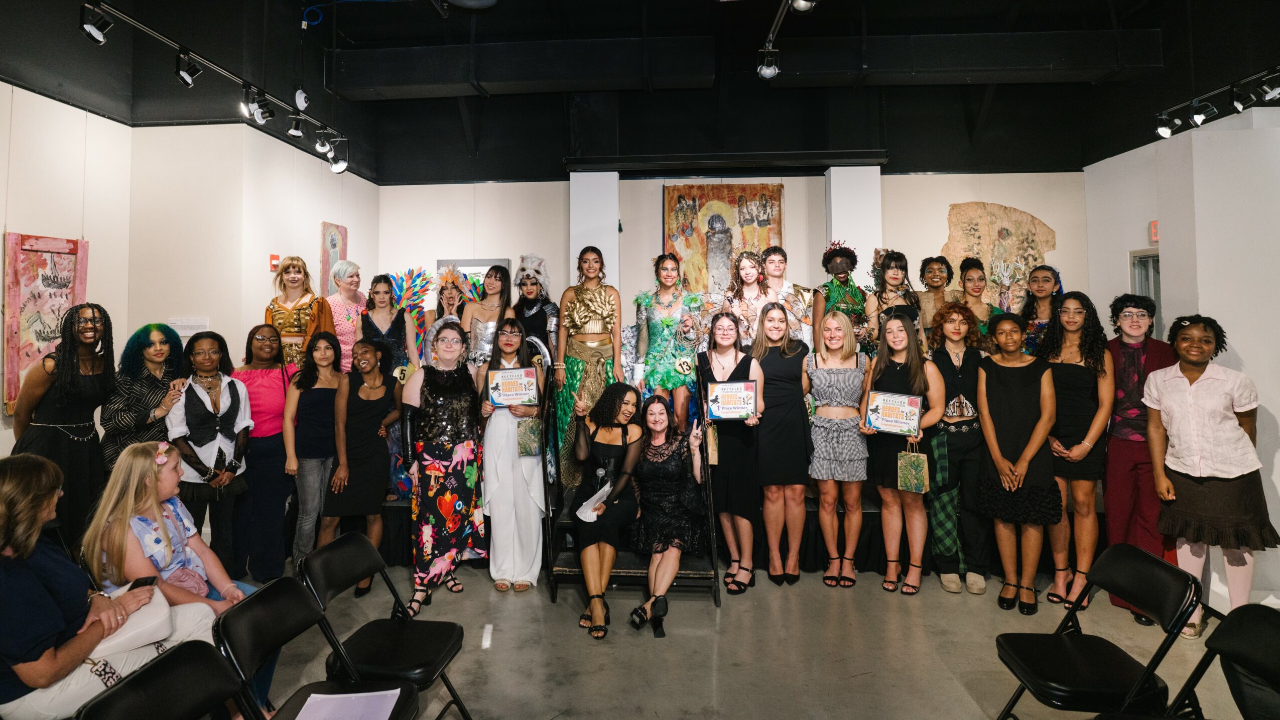 Group of people posing in an art gallery with paintings on white walls, some seated, some standing, diverse attire.