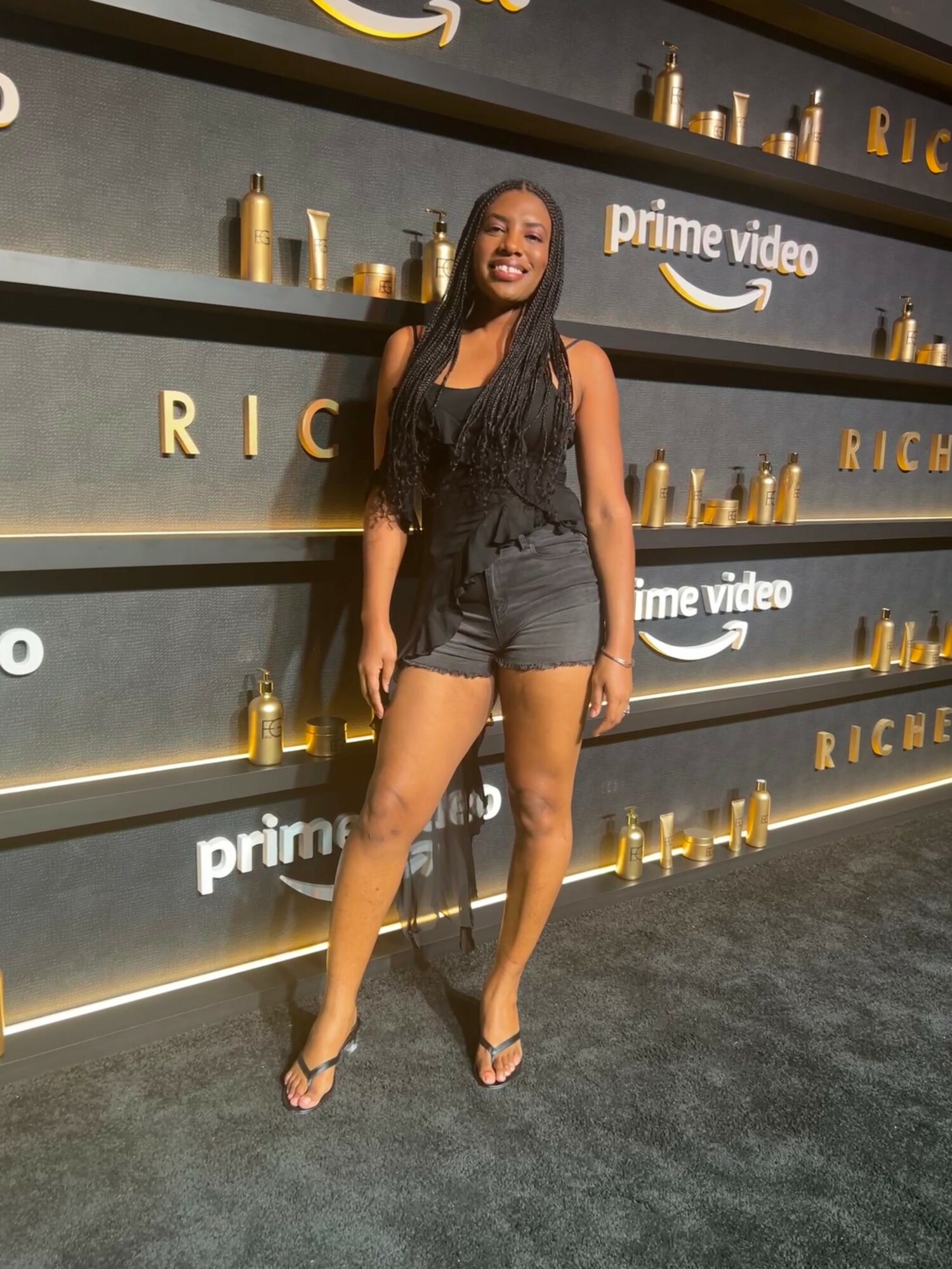 Woman in black outfit standing in front of shelves with gold bottles and Amazon Prime Video logo.