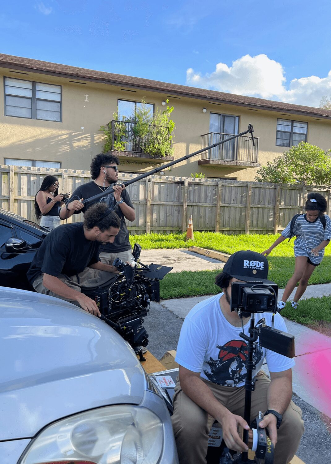 Group of people filming outdoors near a house with a wooden fence, some children playing, camera operator seated with equipment.