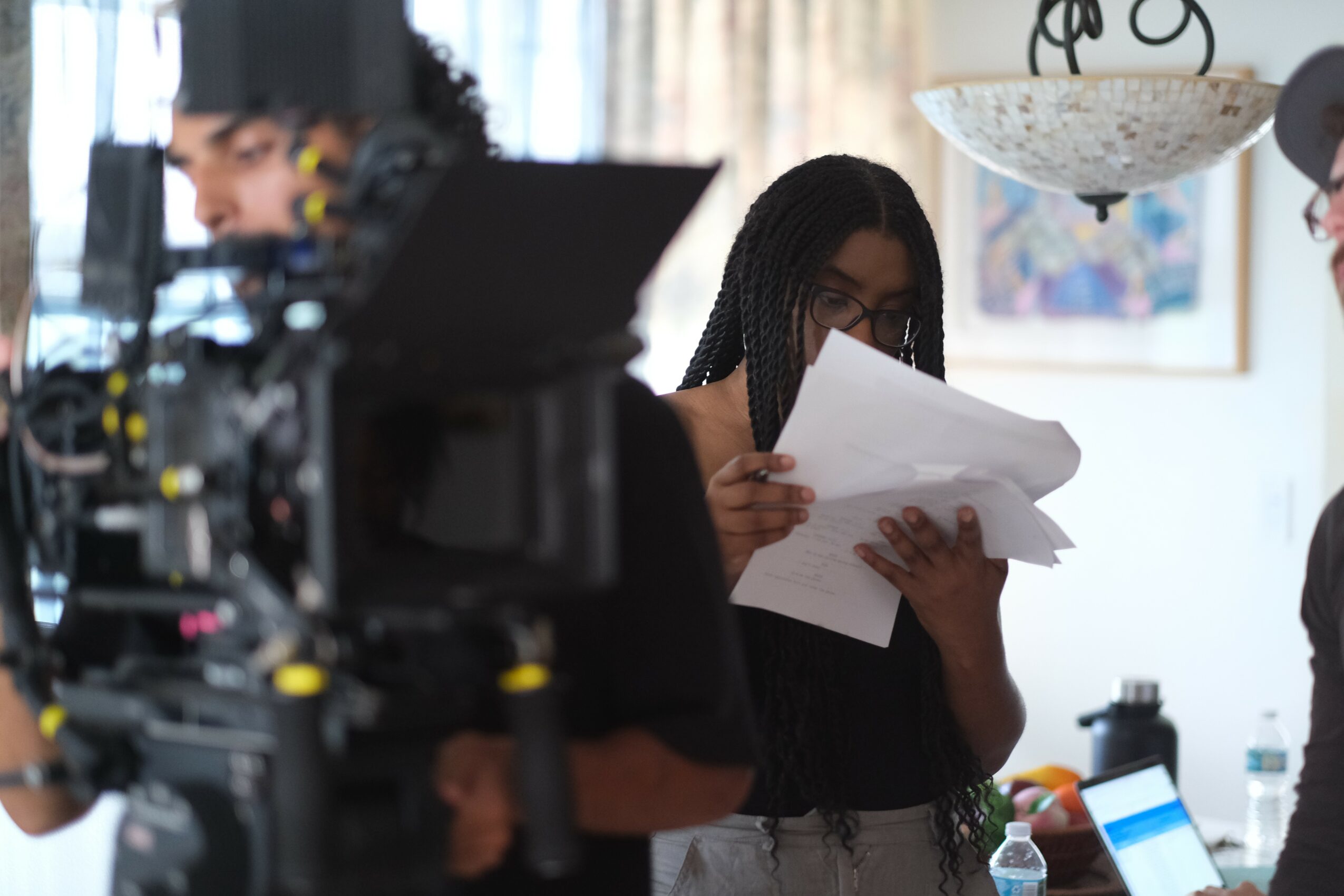 Person with long braided hair and glasses holding papers indoors, with a camera and equipment in the foreground.