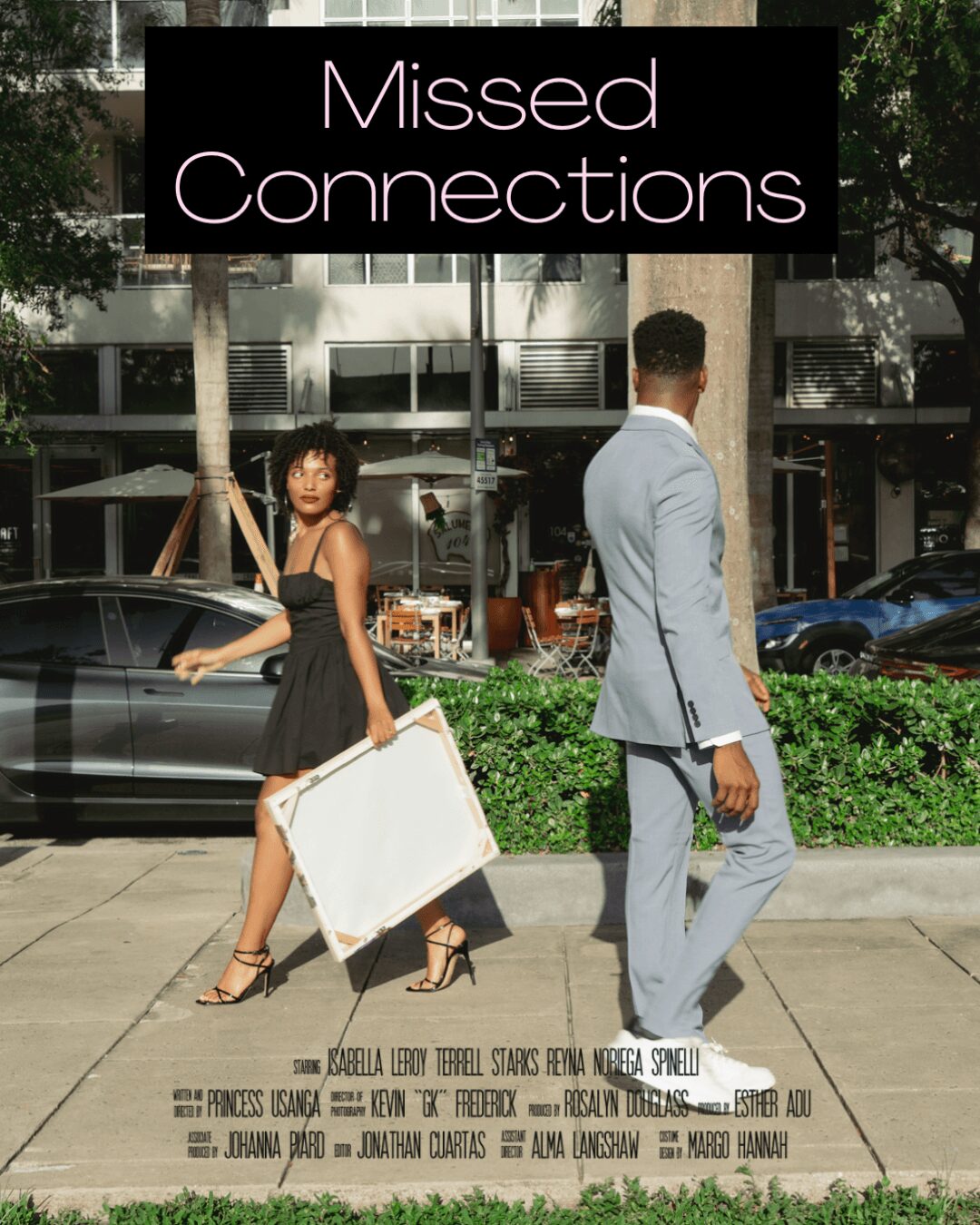Two women walking on sidewalk in front of building, one holding a large white bag, trees and parked cars nearby.