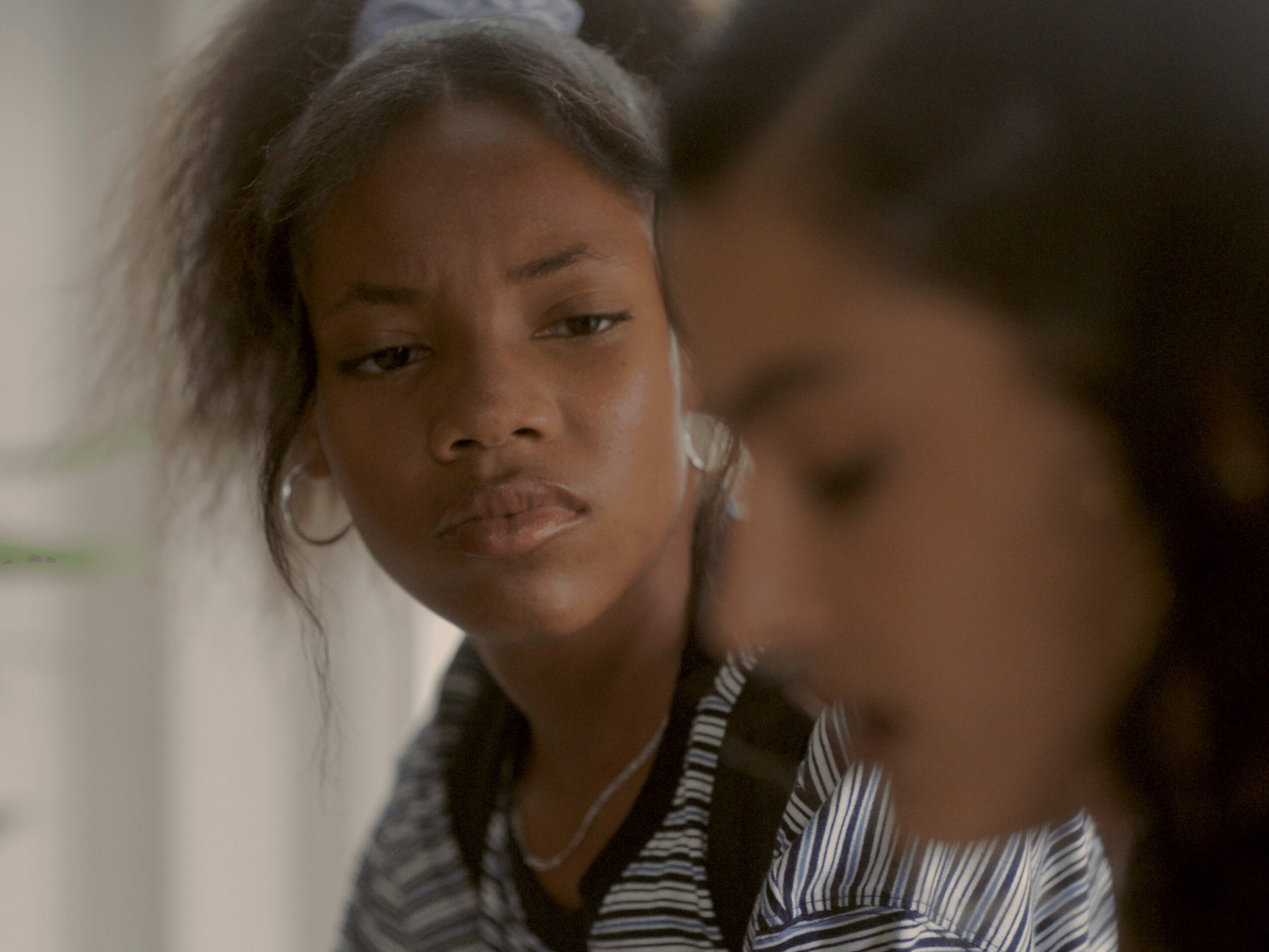 Two women with one in focus and the other blurred, indoors, serious expressions, striped clothing, dark hair, natural lighting.