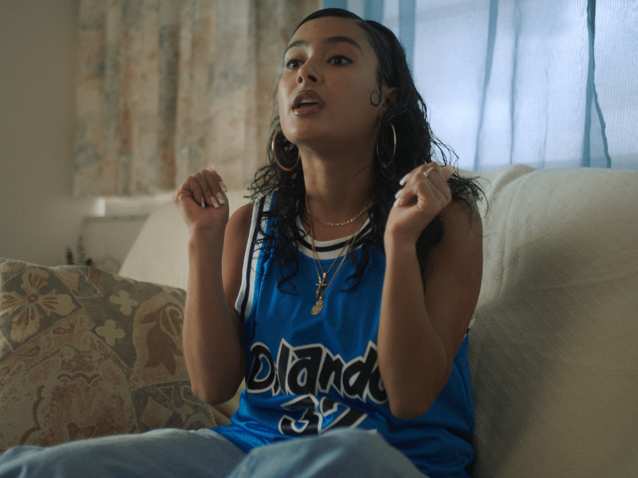Young woman with long dark hair, wearing a blue sports jersey, sitting on a sofa indoors, gesturing with hands.