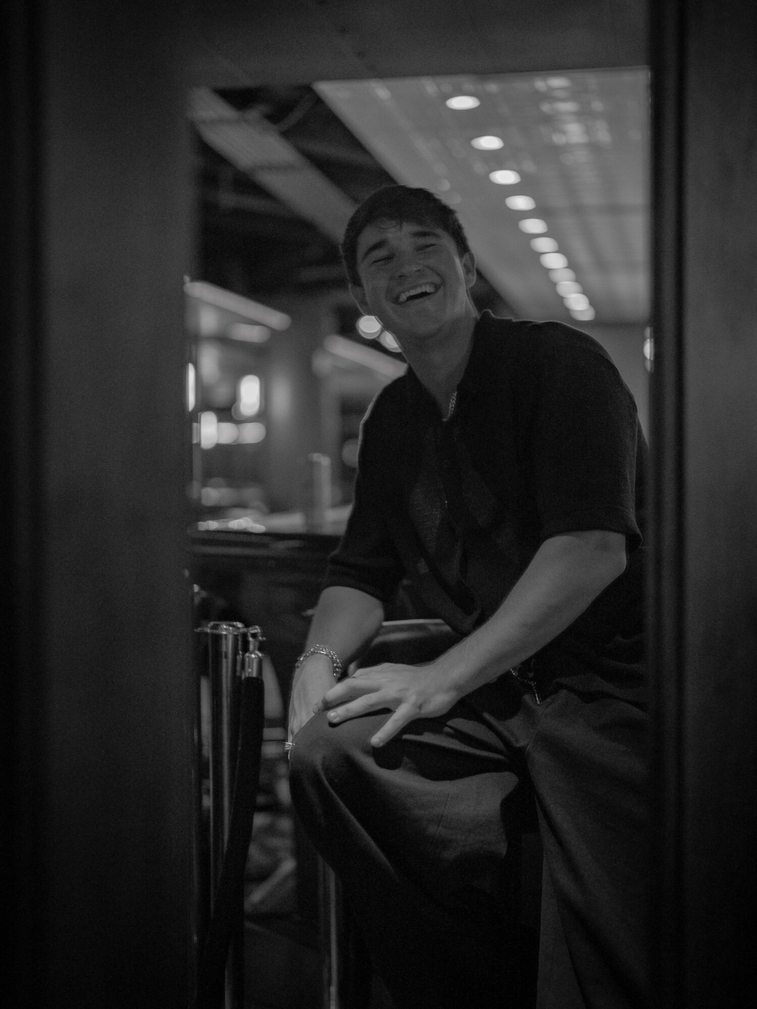 Person smiling and sitting indoors, visible through a window or opening, with ceiling lights in background.