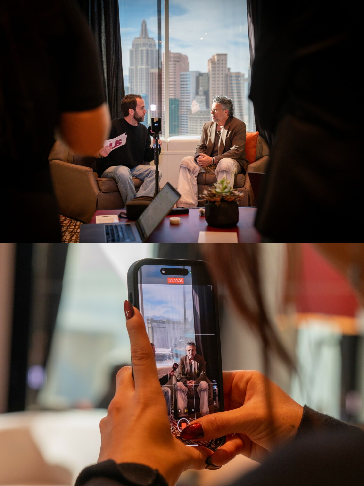 Two people sitting and talking in a modern office with city skyline view, surrounded by colleagues, with a laptop and plant on table.