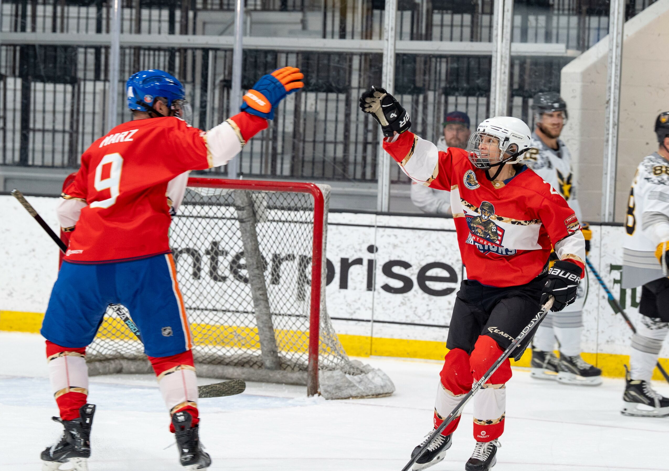 Two ice hockey players in red jerseys celebrate near the goal, with one raising a glove and the other holding a stick.