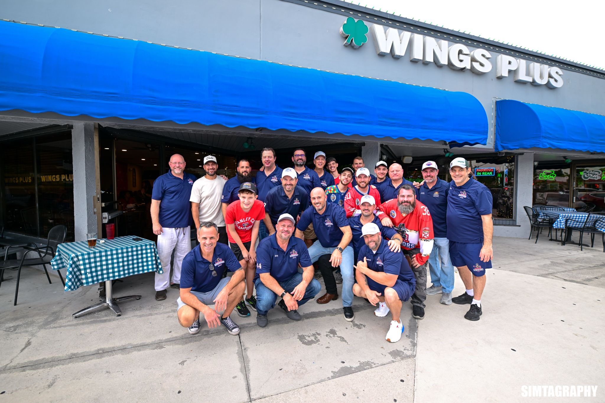 Group of people posing outside a restaurant with a blue awning and a sign reading 'WINGS PLUS'.