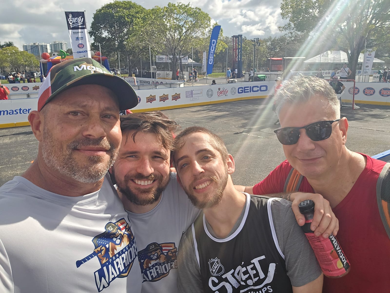 Four men taking a selfie outdoors, some wearing sports jerseys and sunglasses, with a skating rink in the background.