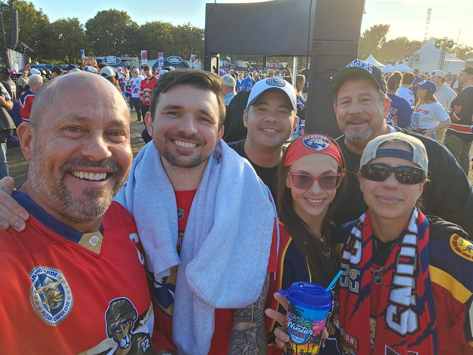 Group of six people smiling outdoors, wearing sports jerseys and accessories, with a crowd and large screen in background.