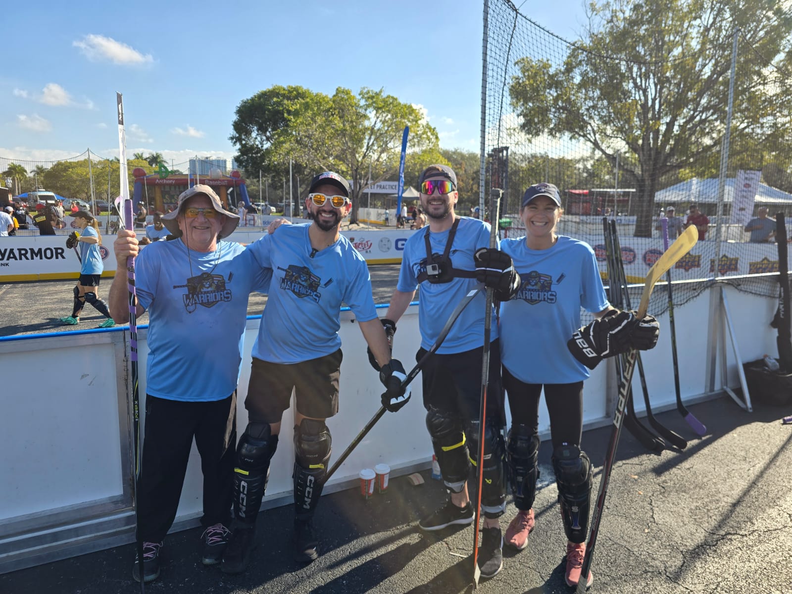 Four people in blue hockey jerseys standing outdoors on a sunny day, holding hockey sticks, near an ice rink barrier.
