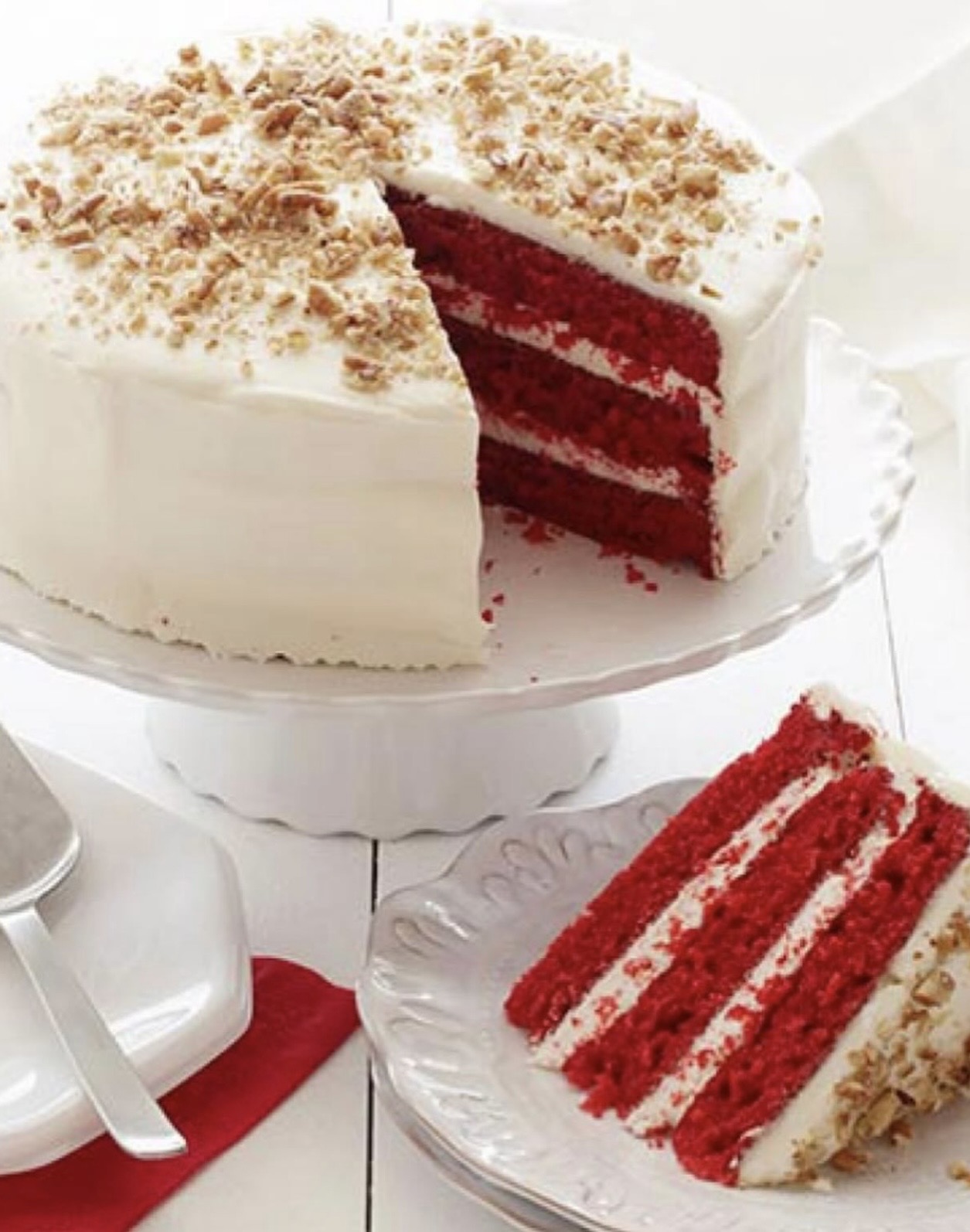 Slice of red velvet cake with cream cheese frosting and crushed nuts on top, on a glass plate.