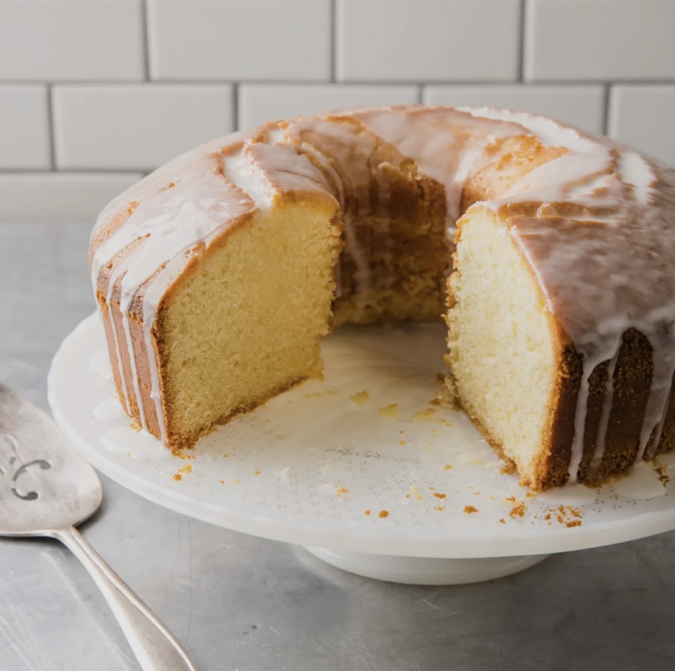 A sliced bundt cake with white glaze on a white plate, on a gray surface with a spoon nearby.
