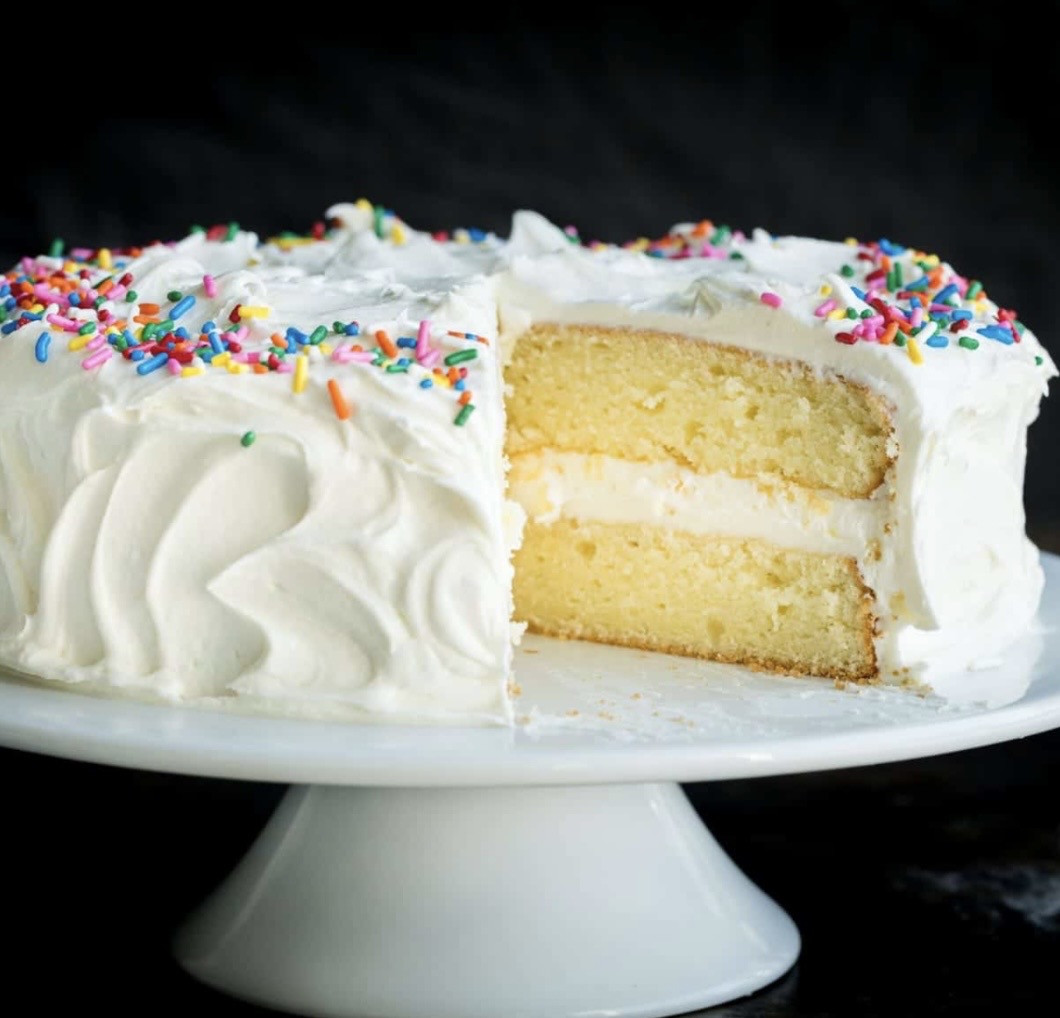 Slice of layered vanilla cake with white frosting and colorful sprinkles on top, on a white cake stand.